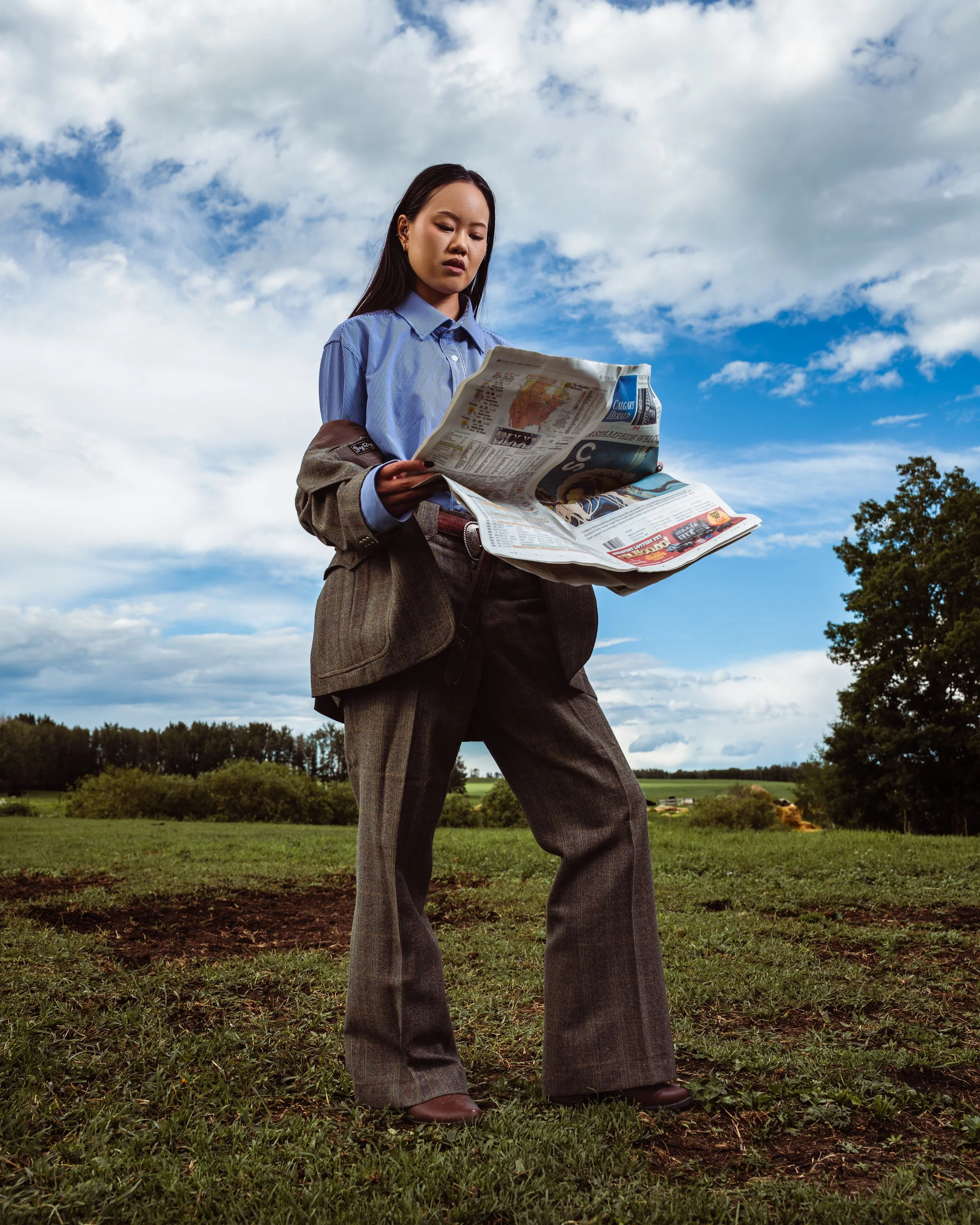 Woman in business clothes reading a newspaper outdoors, with a cloudy sky and trees in the background. Fashion portraits in Cochrane, Alberta by Canadian James Photography.