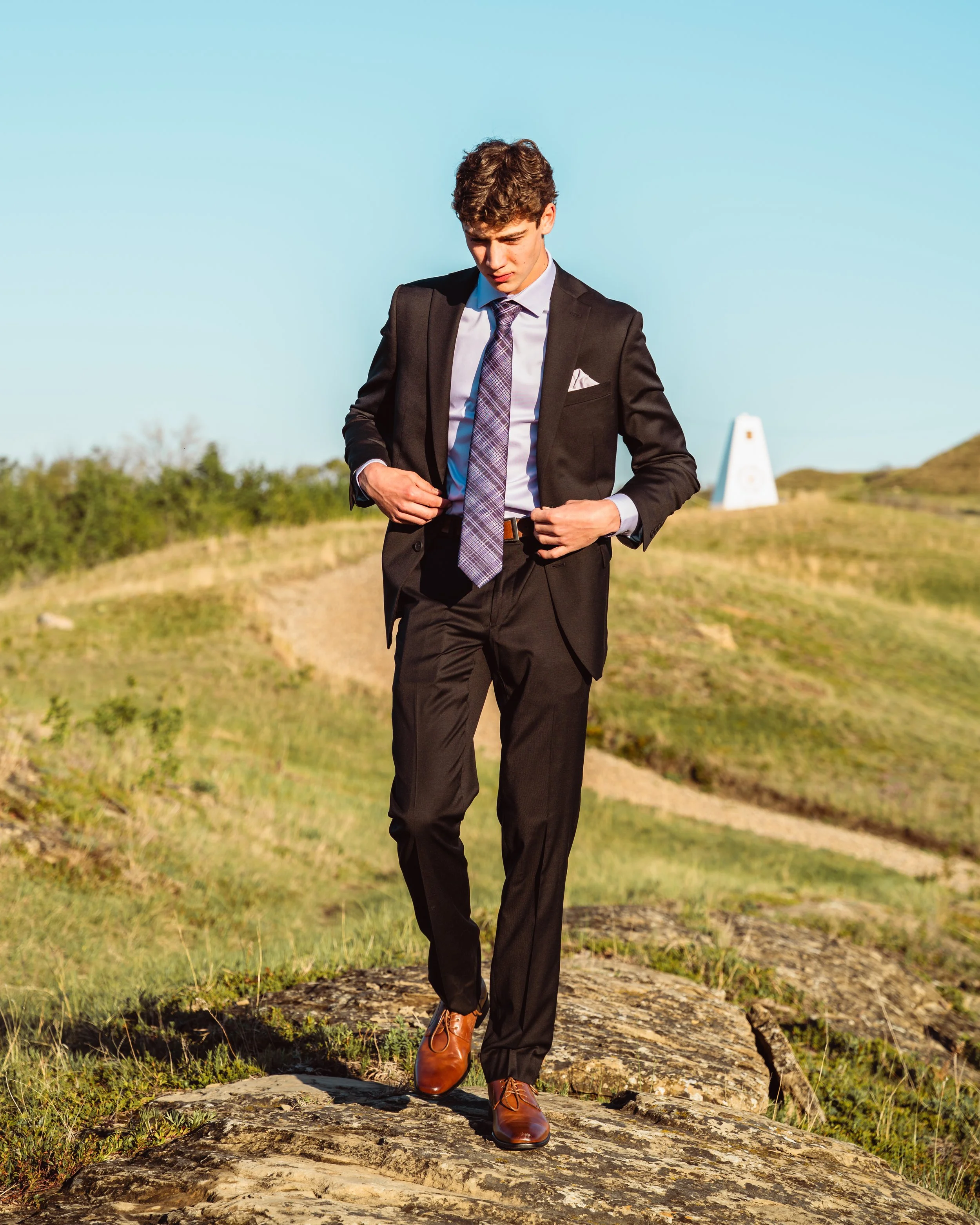 Young man in a suit walking outdoors on a dirt trail, adjusting his jacket, with blue sky and greenery in the background.