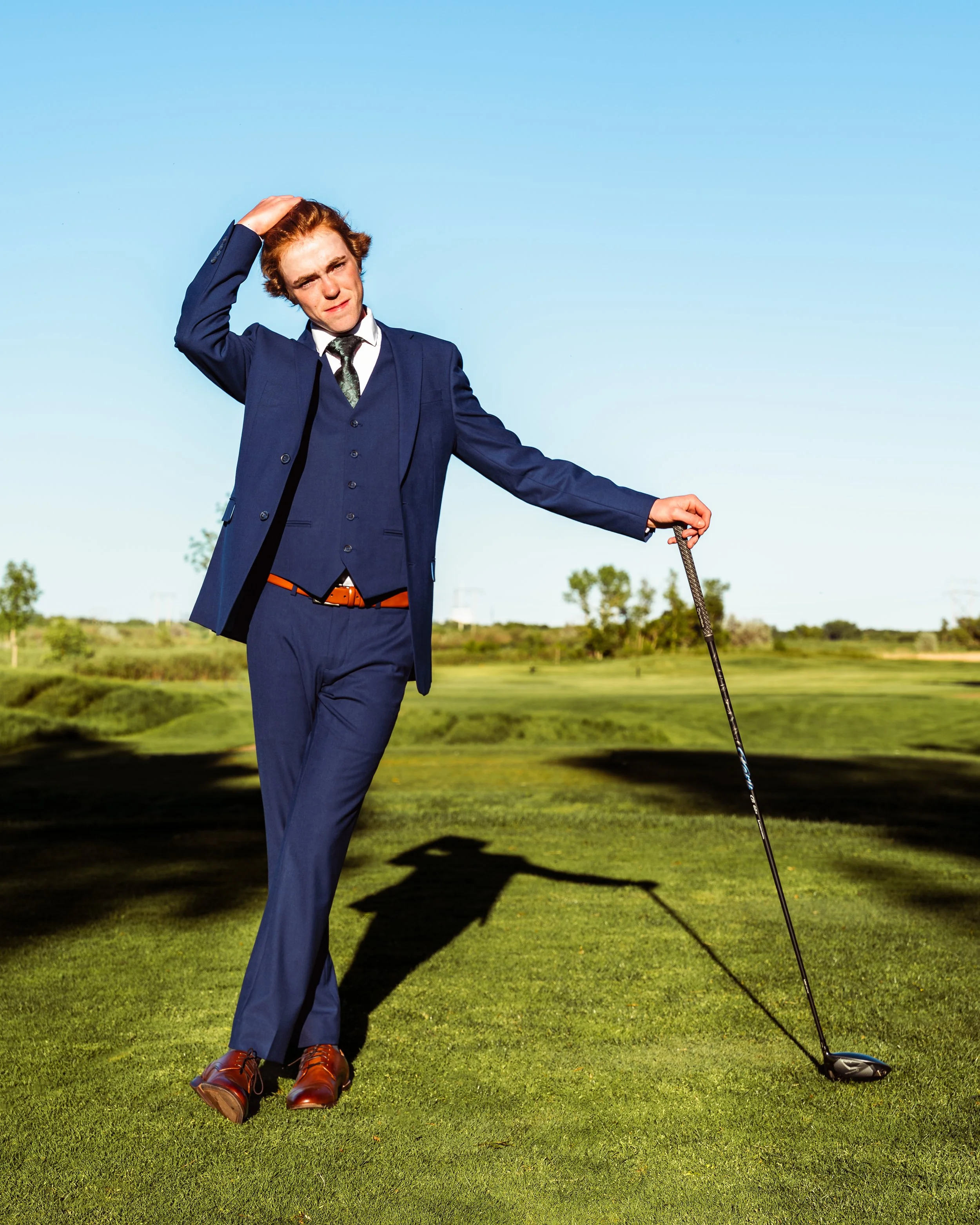 Man in a blue suit with a patterned tie and brown shoes standing on a golf course, scratching his head with his right hand, holding a golf club with his left hand, beneath a clear blue sky. By Canadian James Photography in Calgary, Alberta.