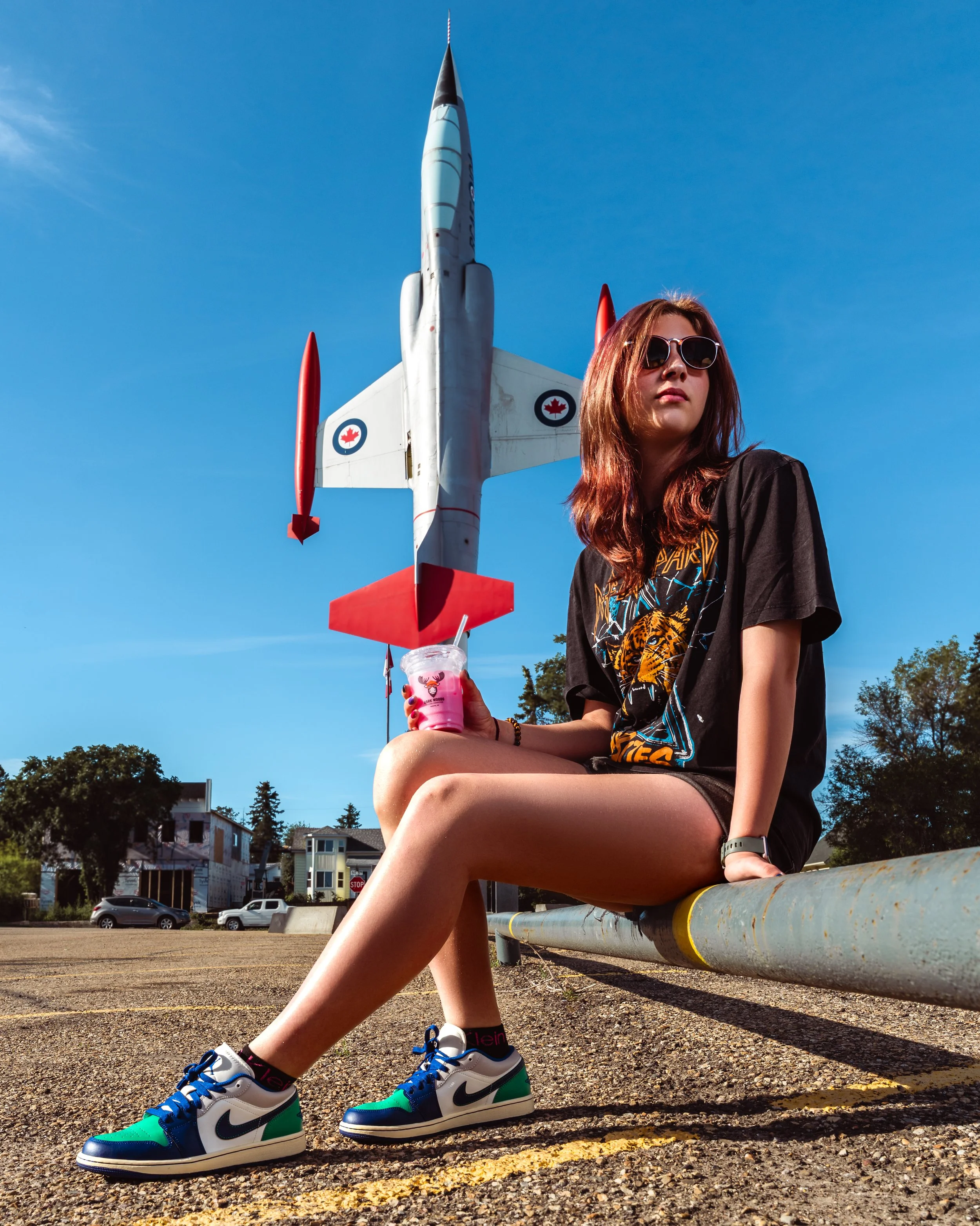 A young woman with red hair wearing sunglasses, a black graphic t-shirt, and multicolored Nike sneakers sitting on a yellow-painted metal rail outdoors. Fashion portraits in Cochrane, Alberta by Canadian James Photography.