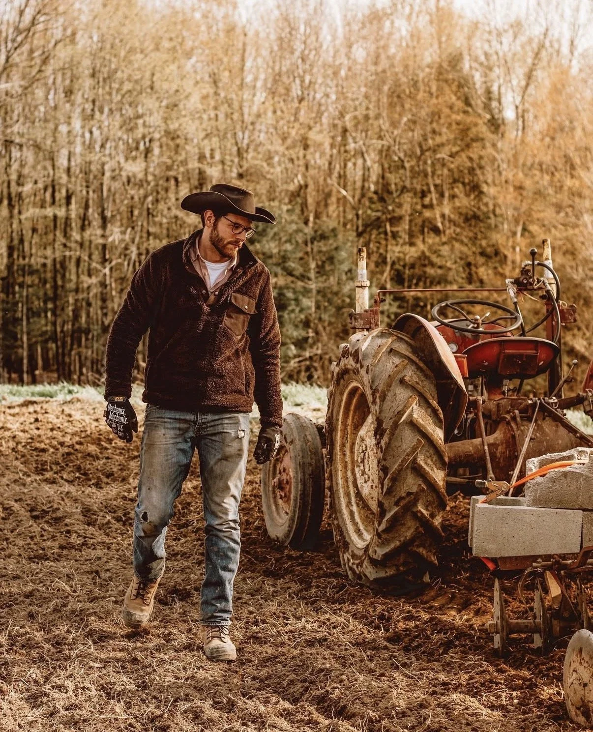A man wearing a black cowboy hat, glasses, a dark jacket, ripped jeans, and work gloves walking through a tilled field beside a rusty tractor on a farm during autumn.