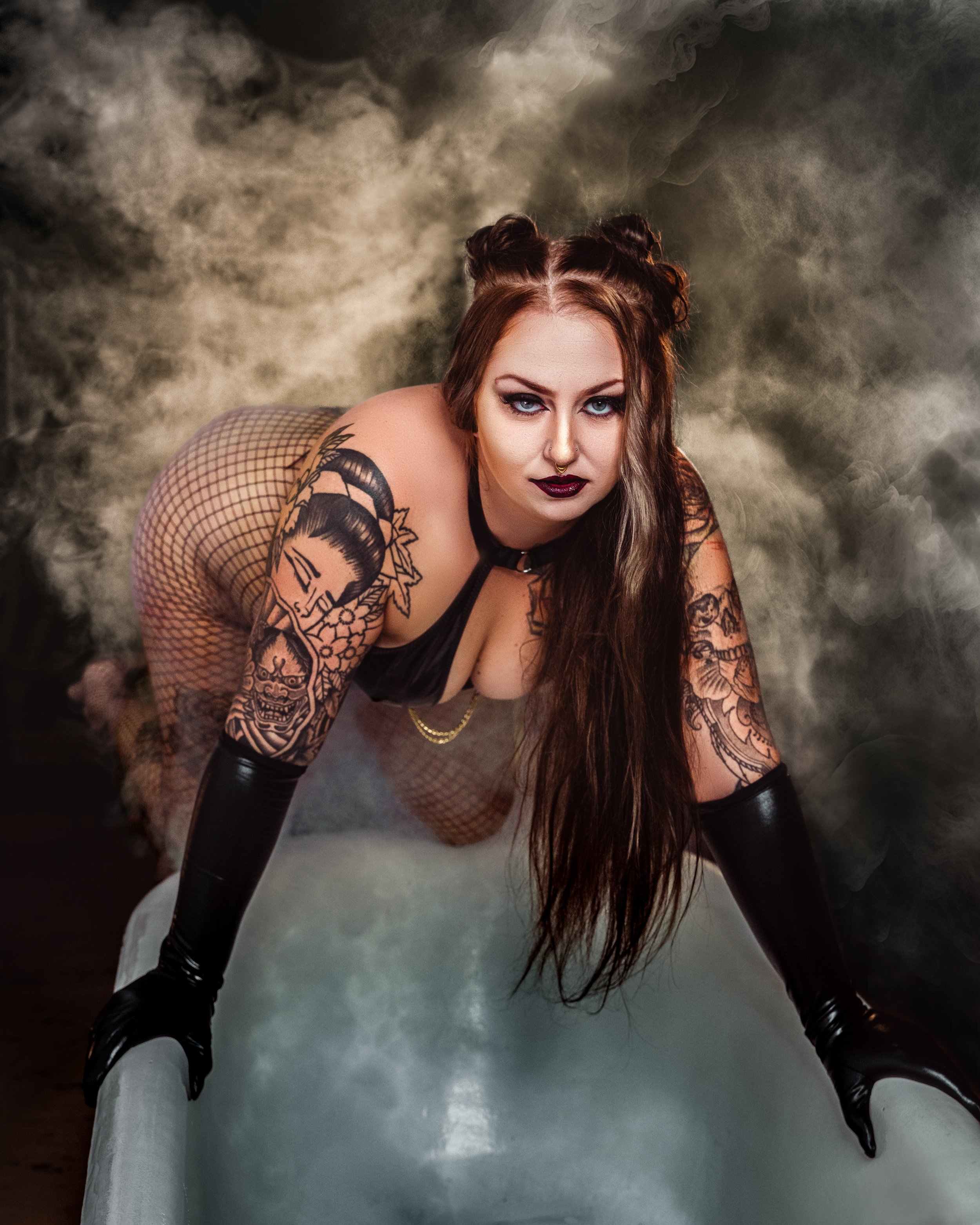 A woman with tattoos and dark makeup crouching with legs apart on a foggy or smoky surface, looking directly at the camera. Cochrane, Alberta photographer.