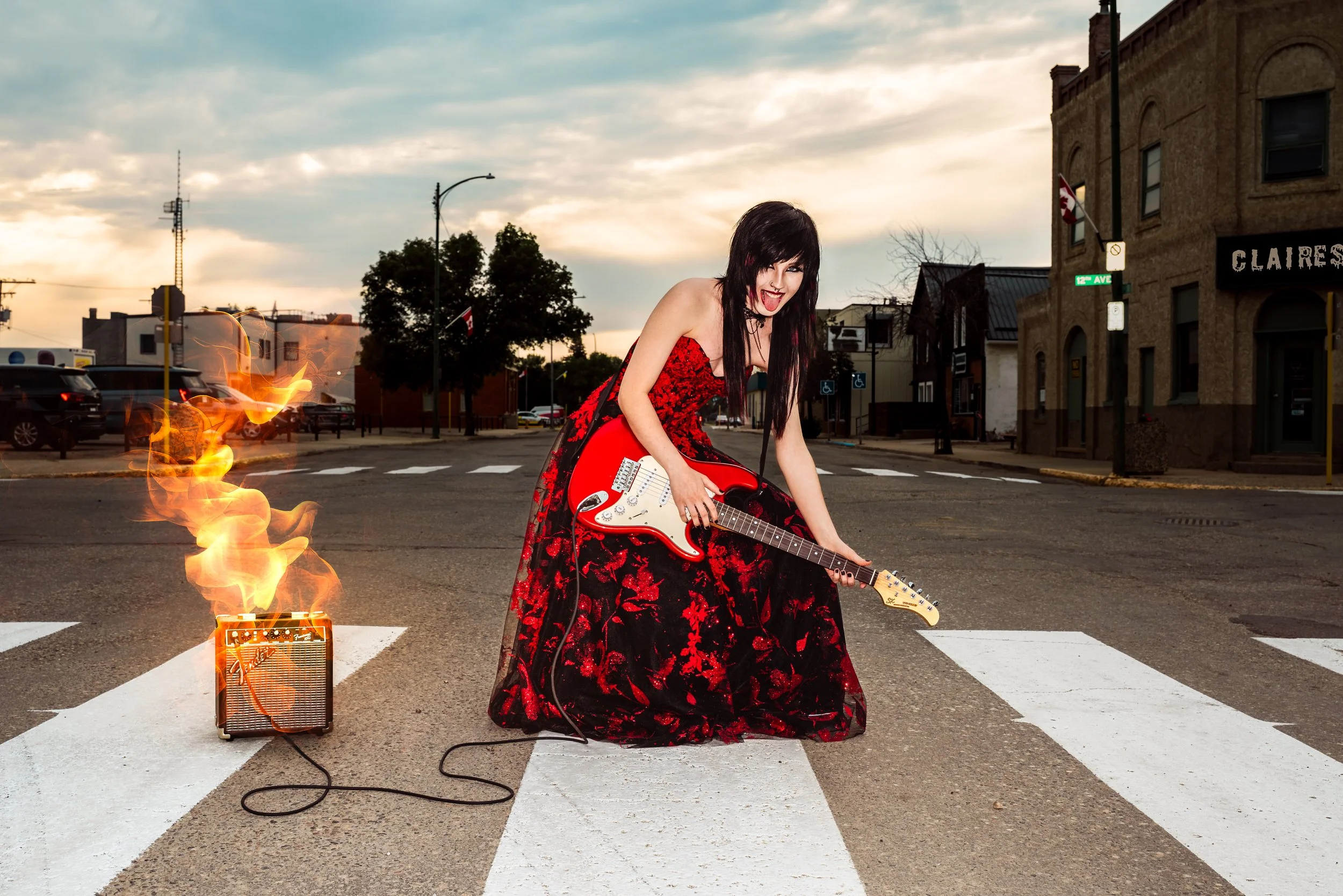 A woman with black hair wearing a red and black floral dress plays an electric guitar on a crosswalk in an urban setting during sunset. Flames and smoke are coming from a small amplifier on the ground, creating a dramatic effect. By Canadian James