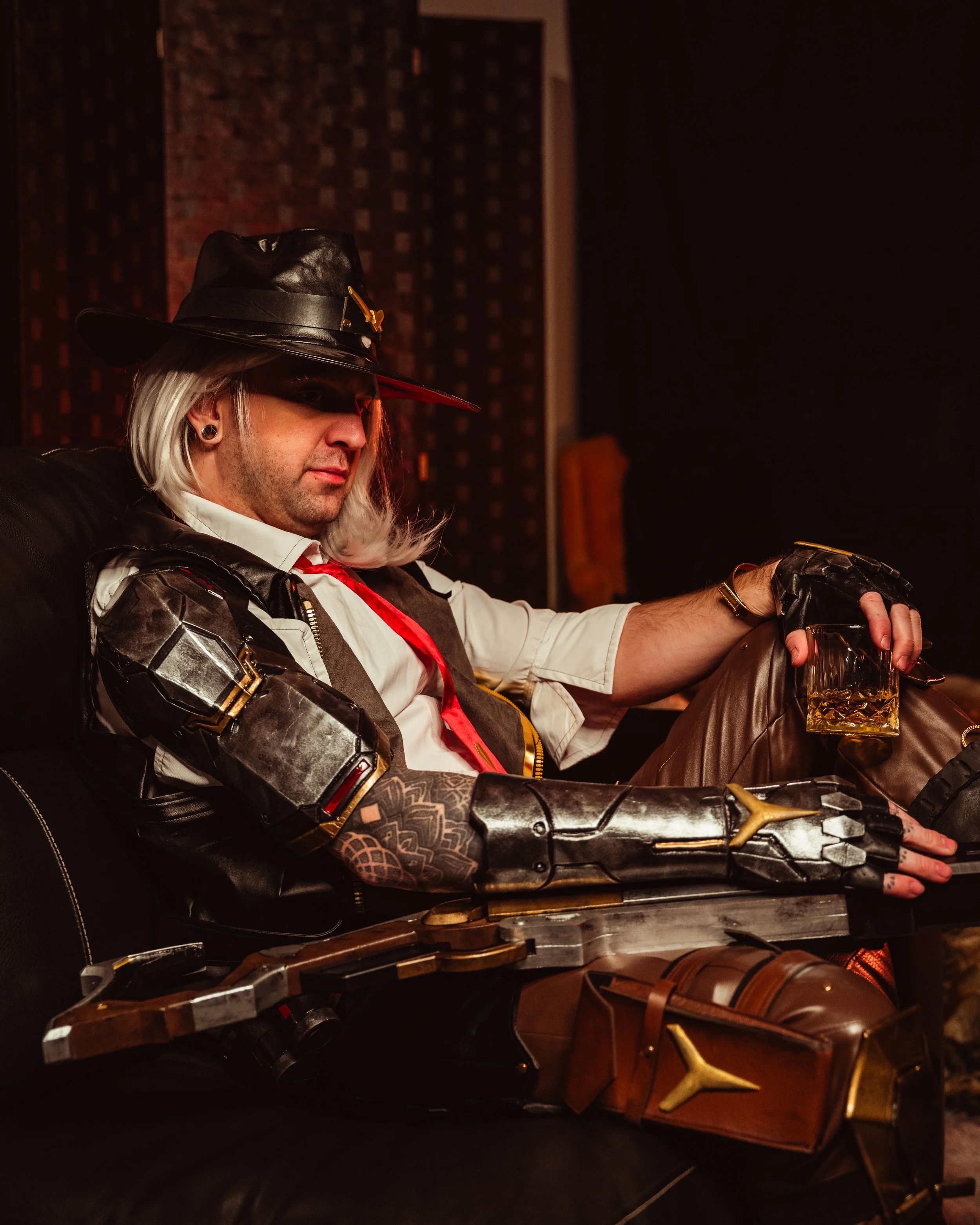 A man dressed in a black leather hat with a gold buckle, a white shirt, and a red tie sits on a black leather chair in a dimly lit room. He has long gray hair, a tattoo on his left forearm, and is holding a glass of whiskey. The room has dark walls with a checkered pattern and faint lighting.