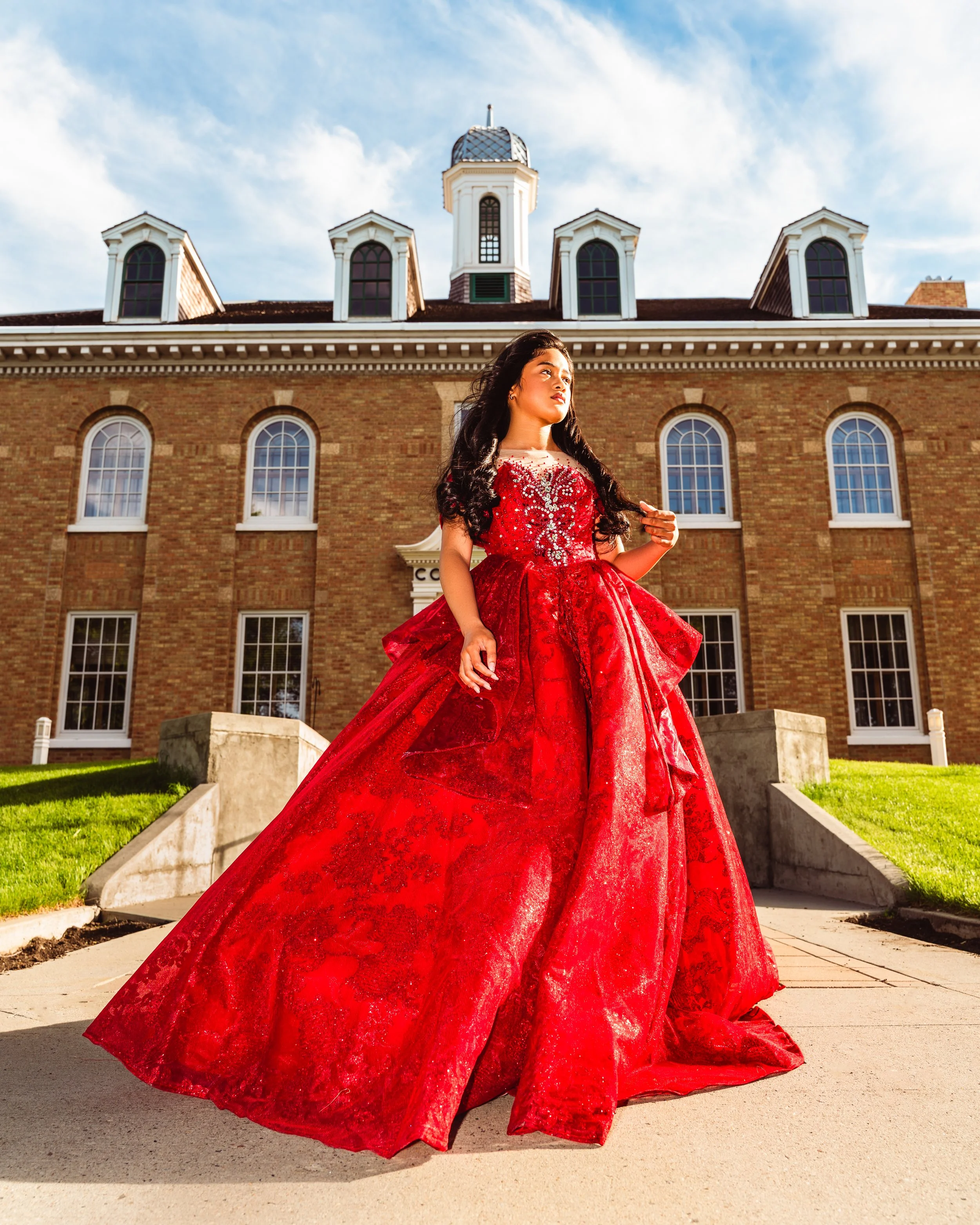 Young woman in a red gown standing outdoors in front of a brick building with multiple windows and a decorative roof, with green grass and a concrete pathway. By Canadian James Photography in Calgary, Alberta.