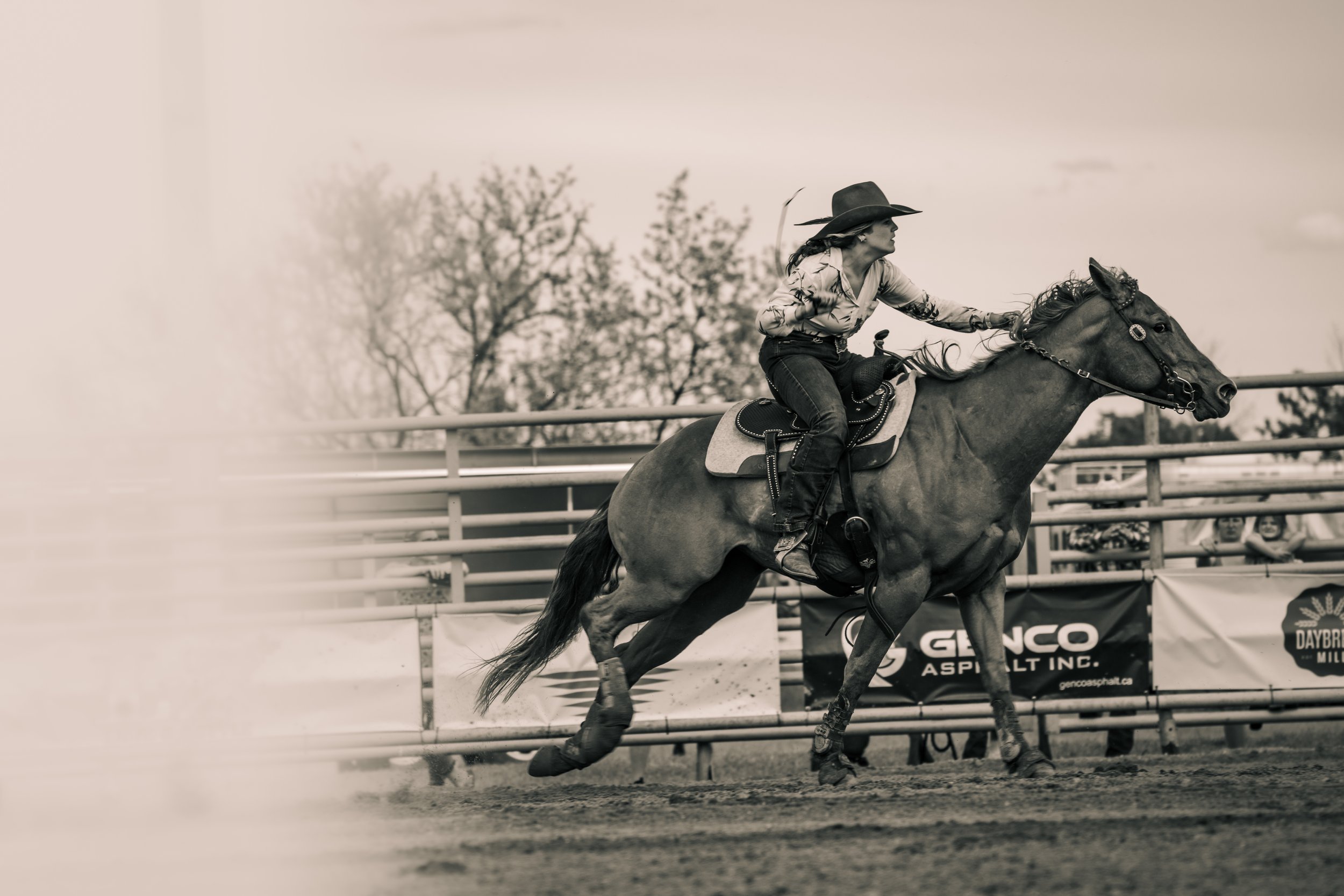 Person in a cowboy hat riding a horse at a rodeo event, galloping fast beside a fence with advertisements. Canadian James Photography. Estevan, Saskatchewan.