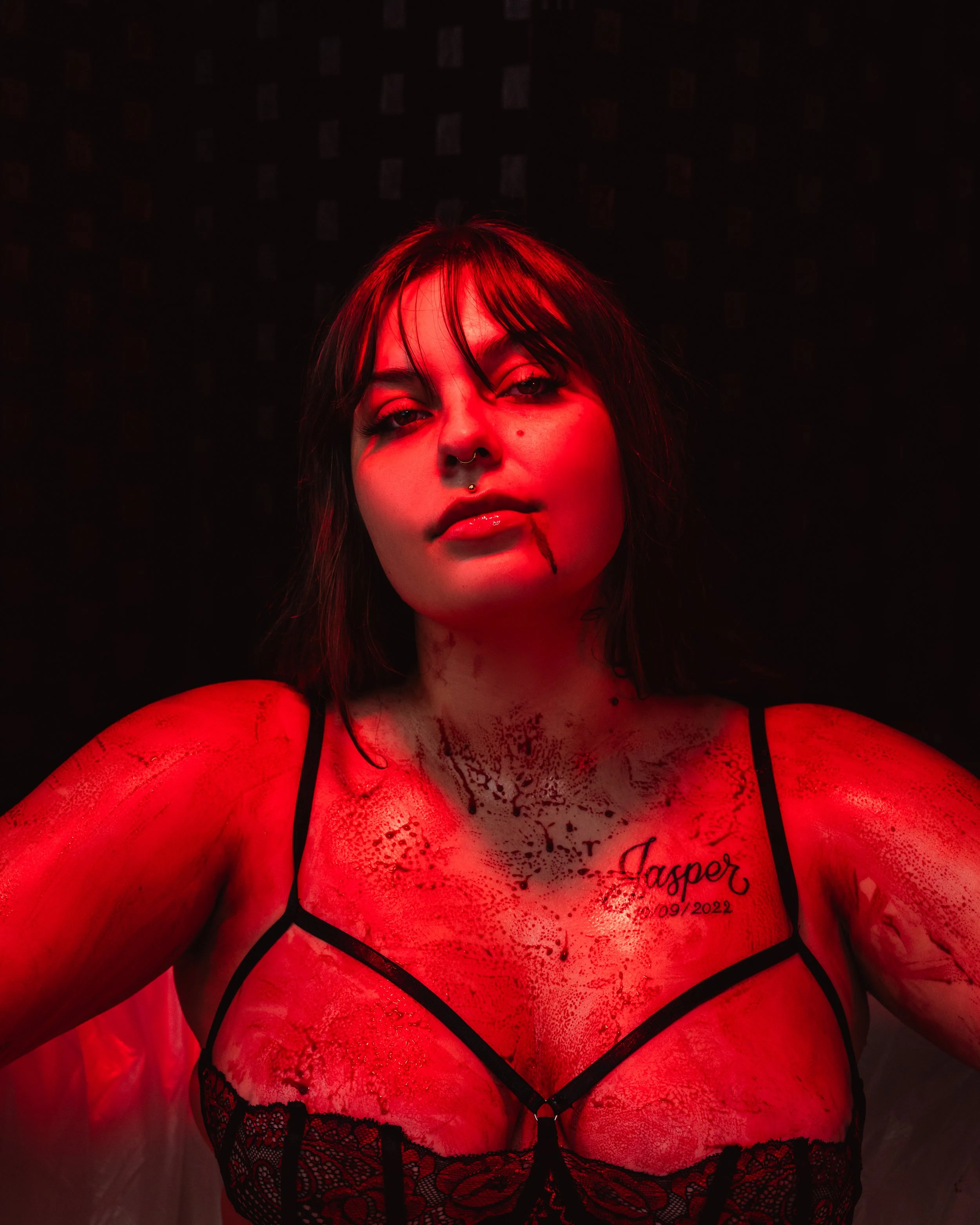 A woman with dark hair and tattoos wearing black lace lingerie, illuminated by red light, dark background.