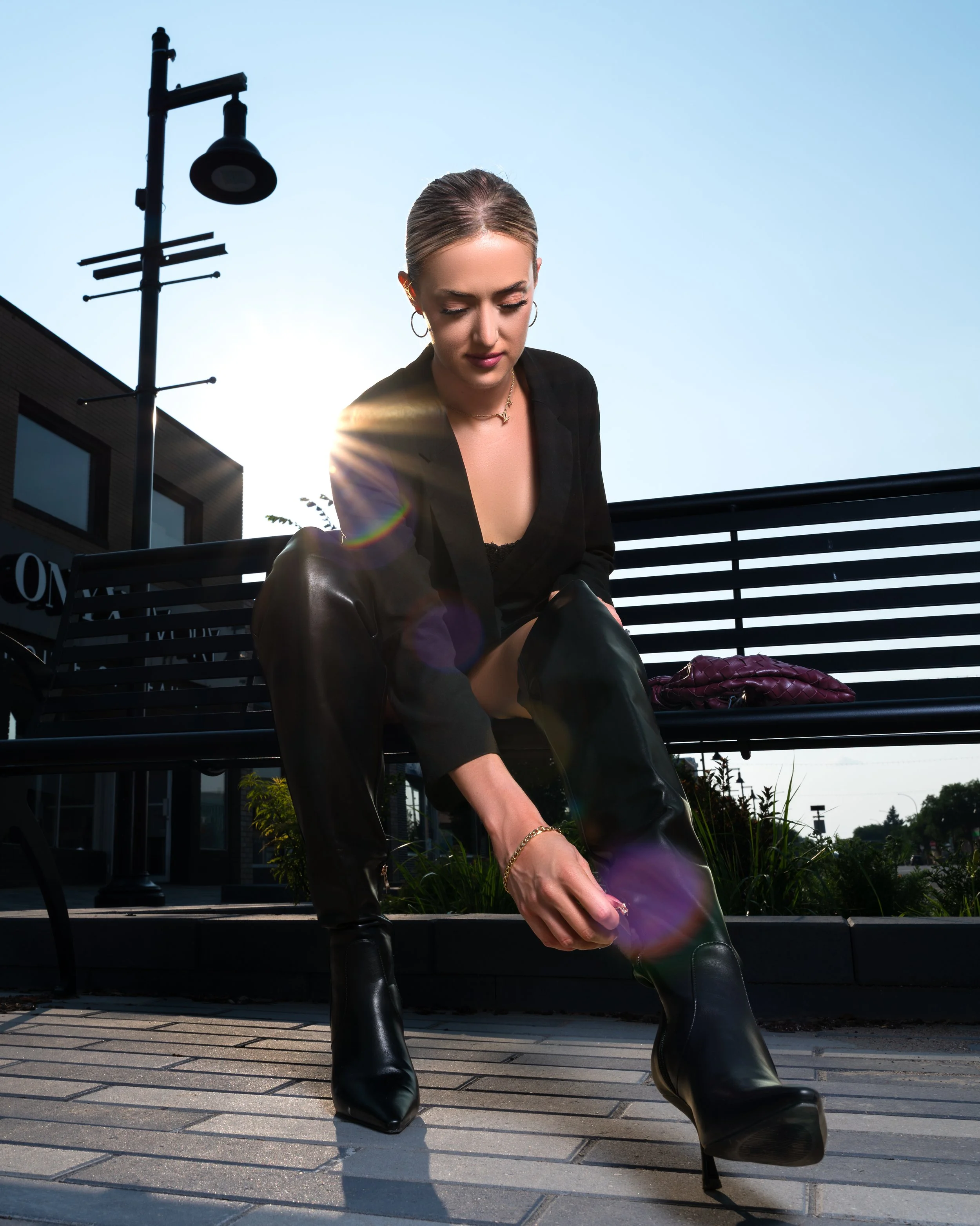 Woman sitting on a bench outdoors, looking down, with sunlight shining behind her and a clear blue sky. Top Calgary Portrait Photographer.