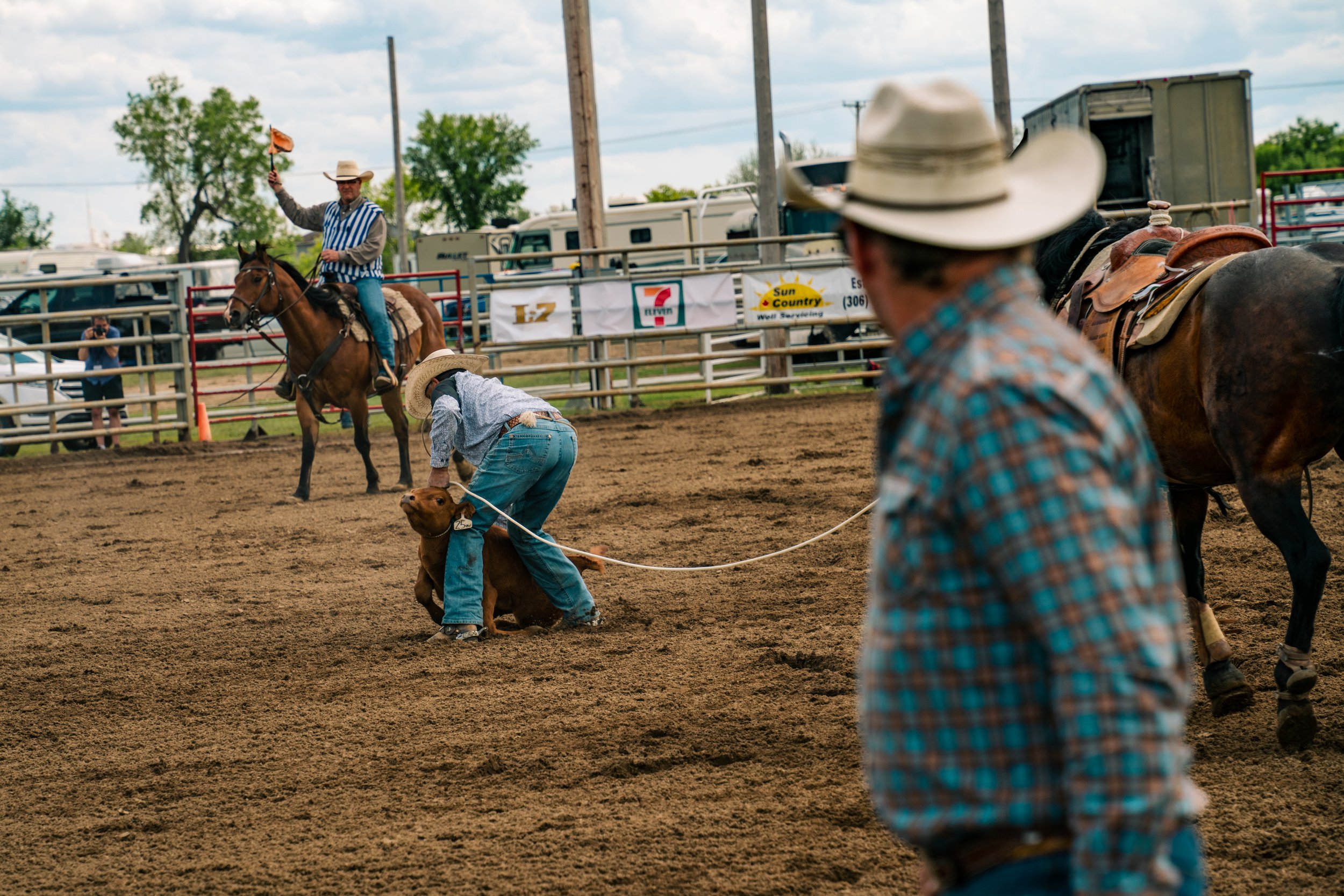 Rodeo scene with cowboy in hat tying a calf, another on horseback, spectators in background.