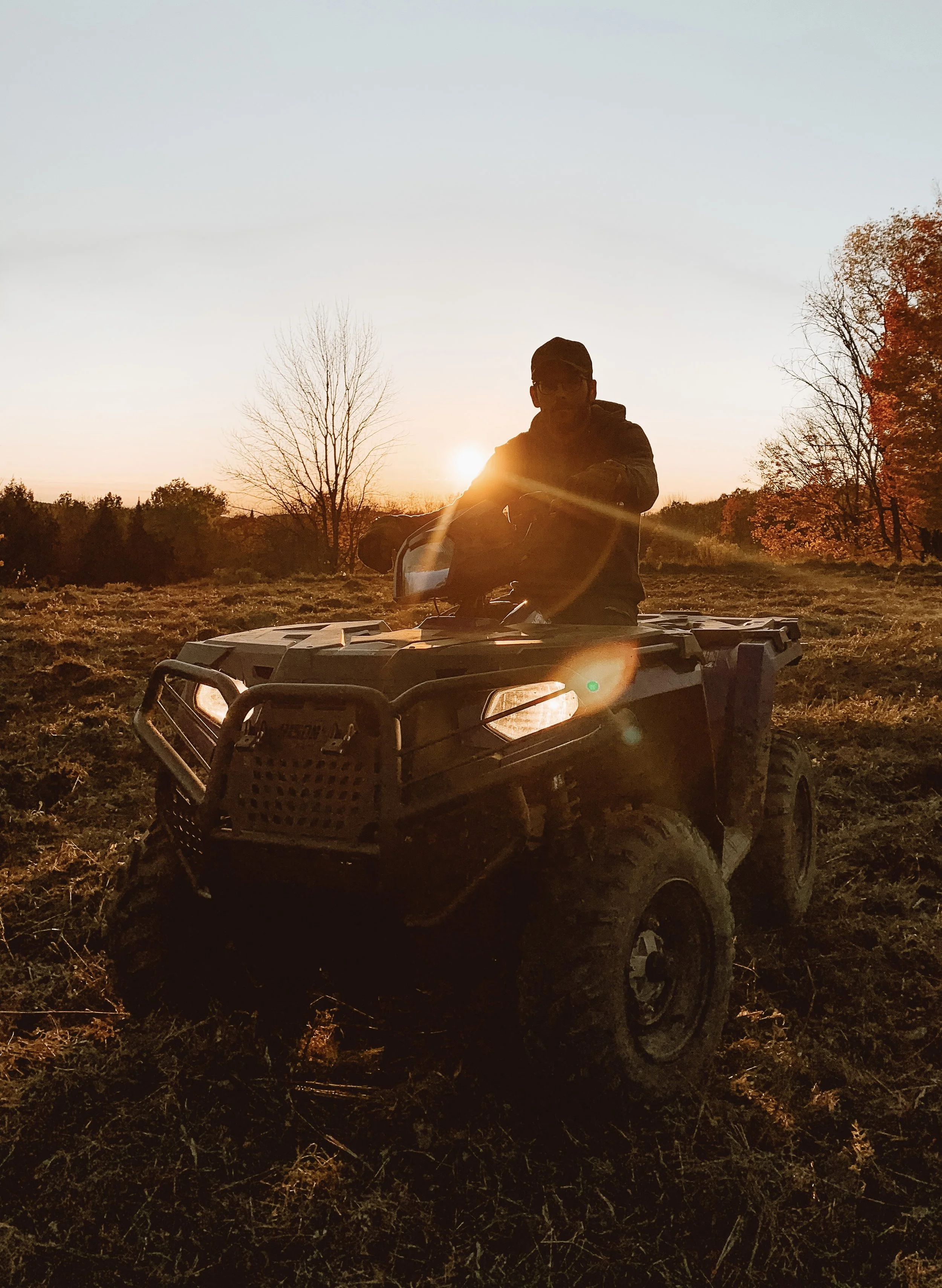 Person riding an all-terrain vehicle (ATV) during sunset in a field with trees.