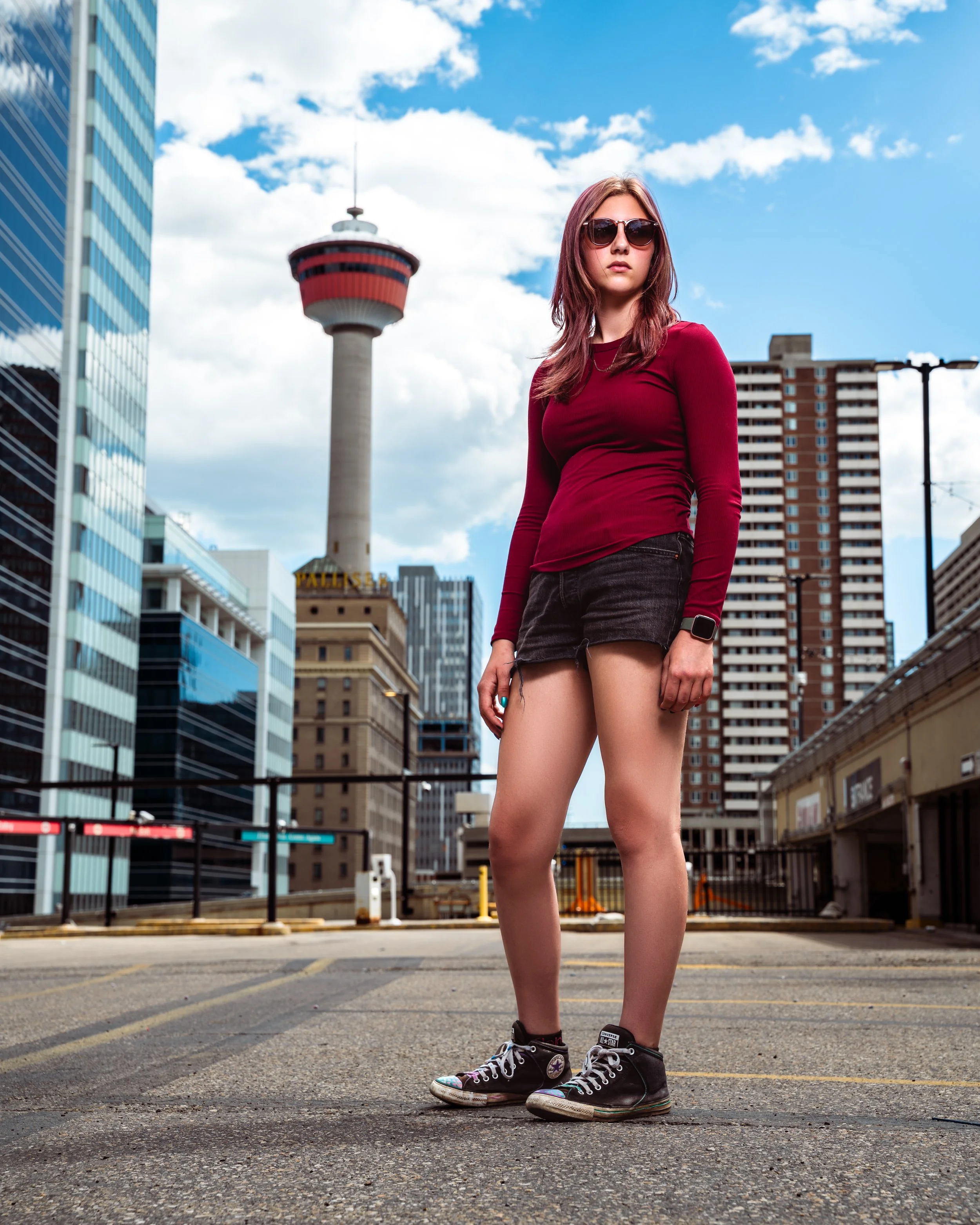 A young woman with brown hair wearing sunglasses, a red long sleeve shirt, black shorts, and black Converse sneakers standing in an urban parking lot with tall modern buildings and the Calgary tower in the background. Calgary, Alberta