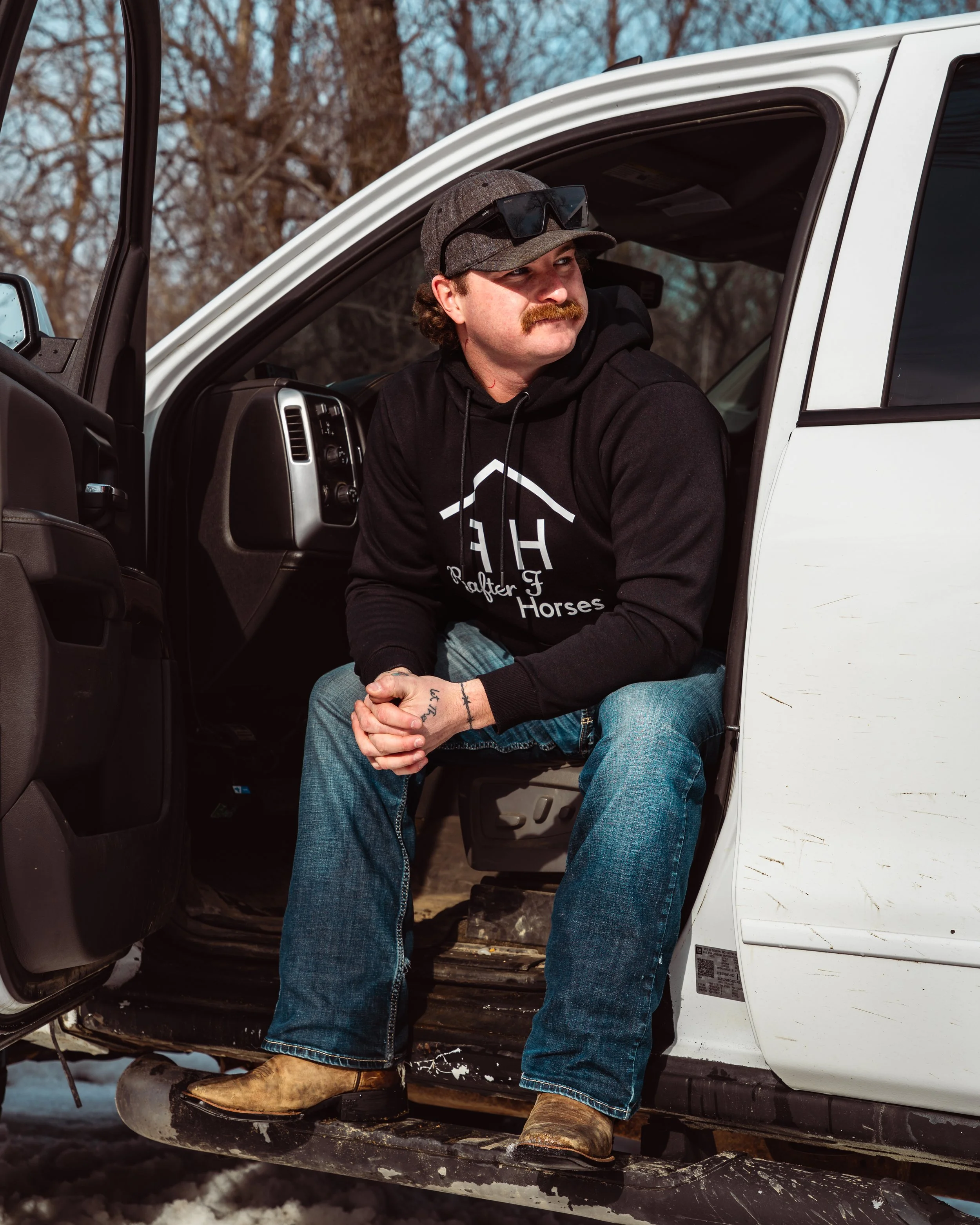 A man with a mustache sitting inside an open door of a white pickup truck, wearing jeans, work boots, a black hoodie, sunglasses, and a cap, with trees in the background.