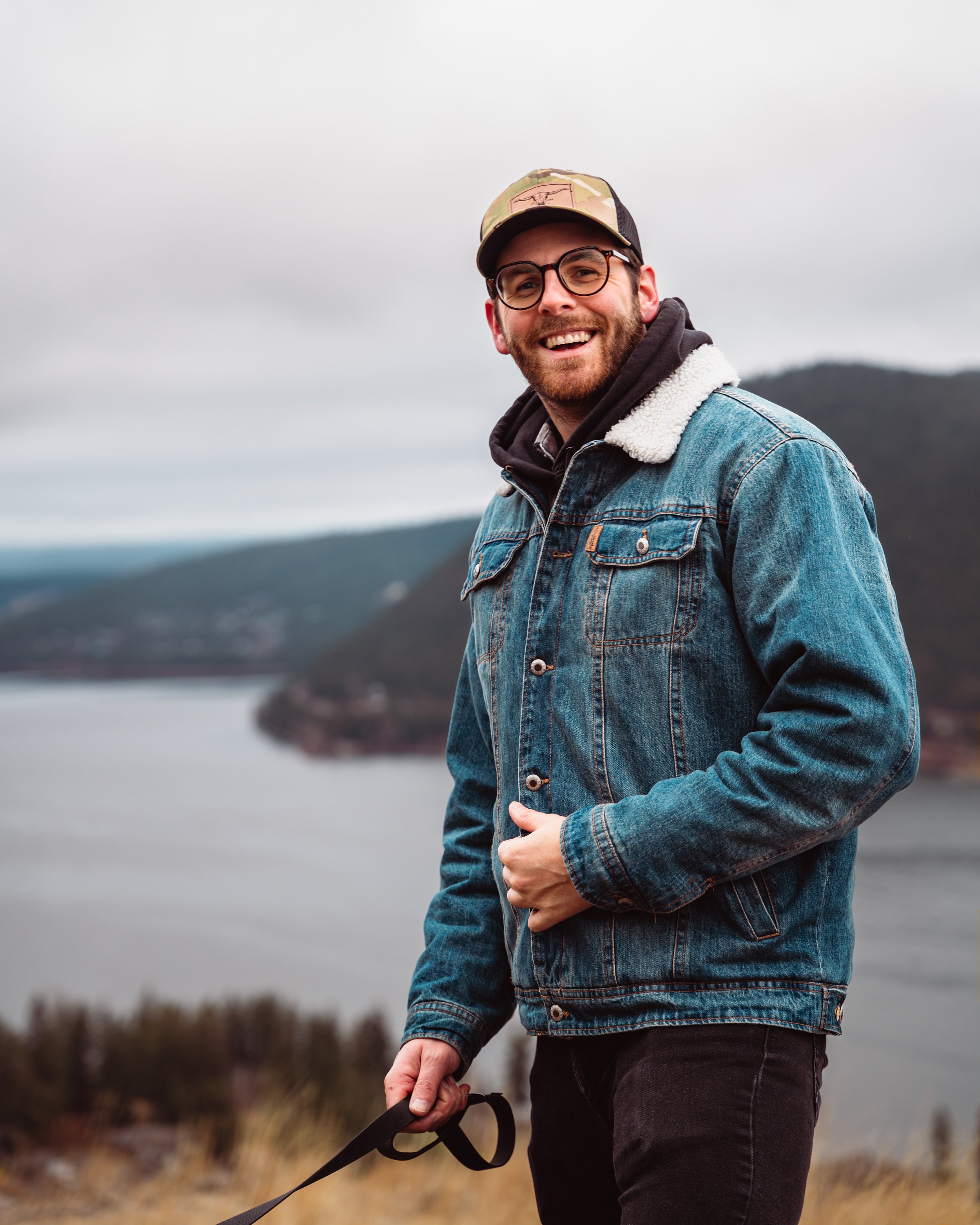 A smiling man in a denim jacket and camouflage cap outdoors, holding a leash, with overcast sky and distant overgrown landscape in the background. Cochrane Alberta photographer.