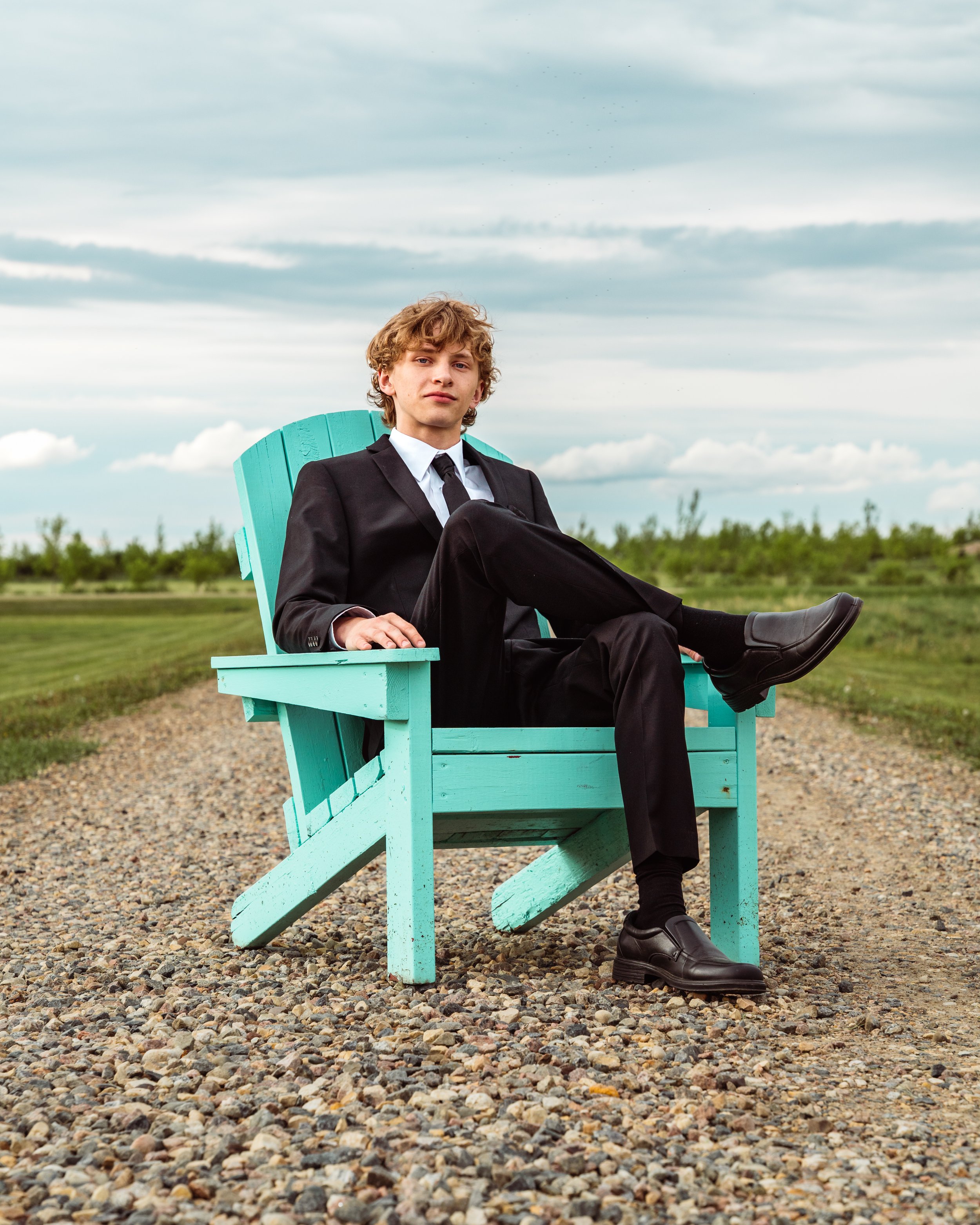 A young man with curly blond hair dressed in a black suit, white shirt, and black tie sitting on a turquoise wooden Adirondack chair outdoors, with a grassy field and cloudy sky in the background. By Canadian James Photography in Calgary, Alberta.