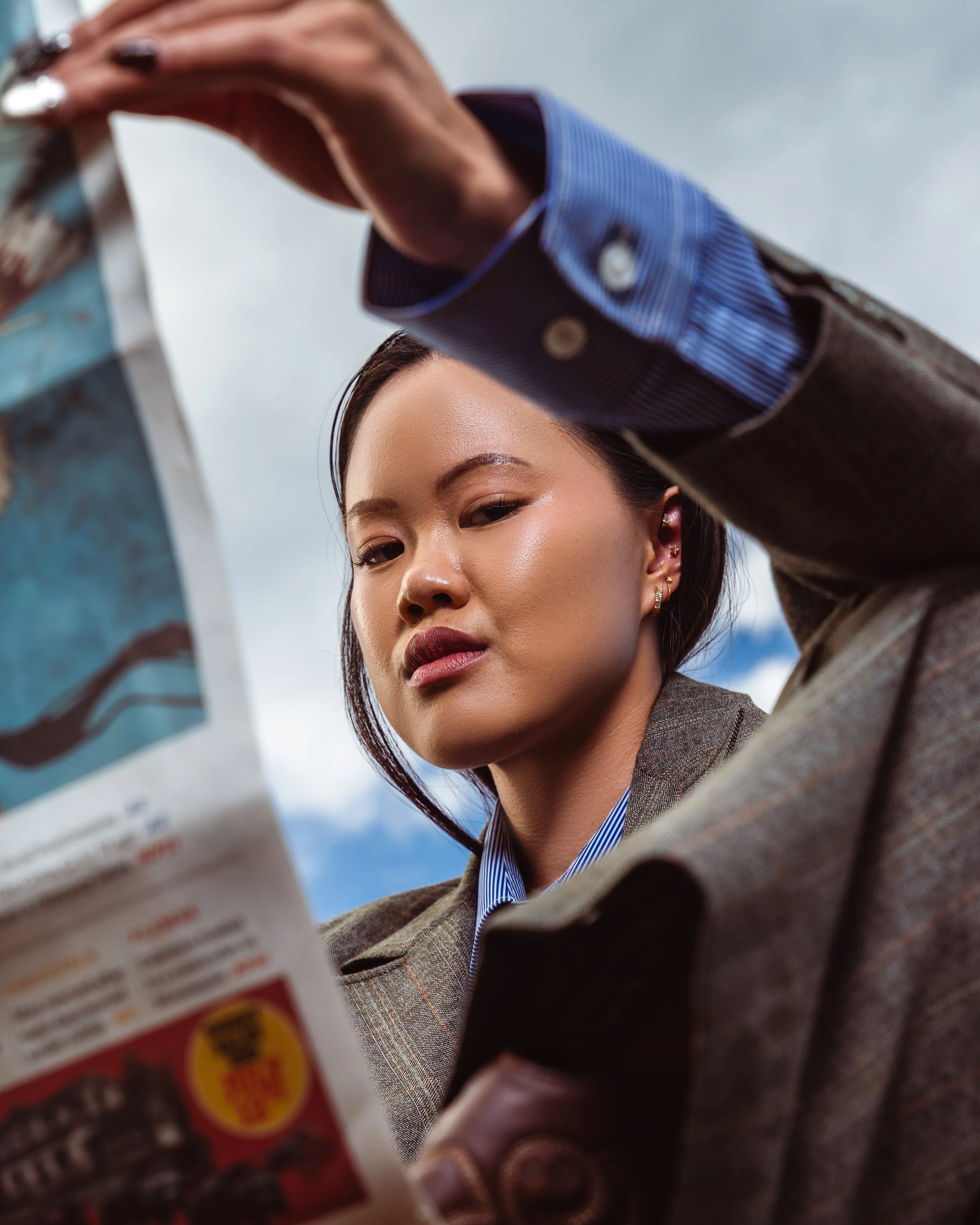 A woman with dark hair, light skin, and pierced ears reading a newspaper outdoors on a cloudy day. Fashion portraits in Cochrane, Alberta by Canadian James Photography.
