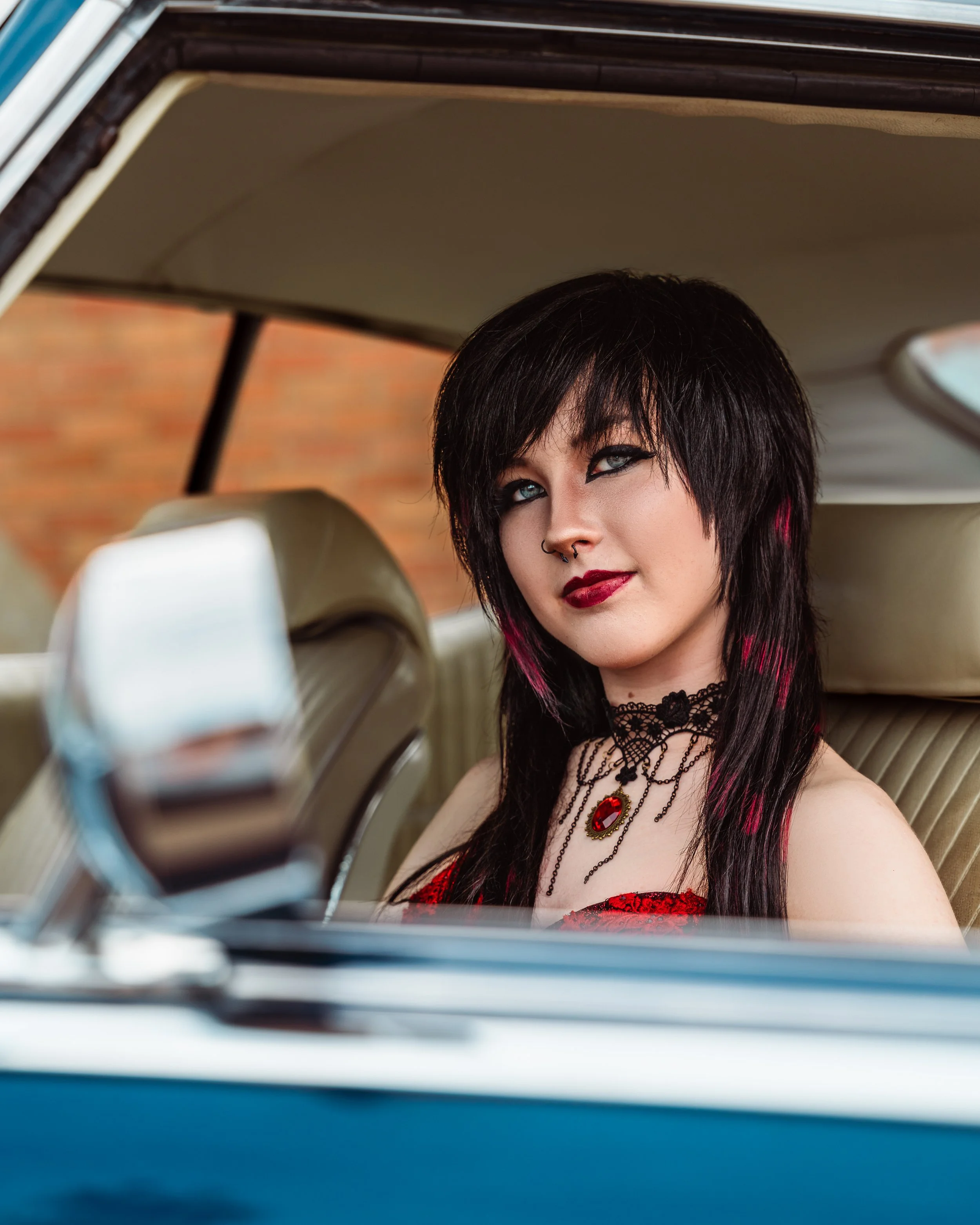 A woman with black hair and blue eyes looking out from the backseat of a car, wearing a red dress with black lace and jewelry, with the photo taken through the car window. Calgary Alberta grad photographer.