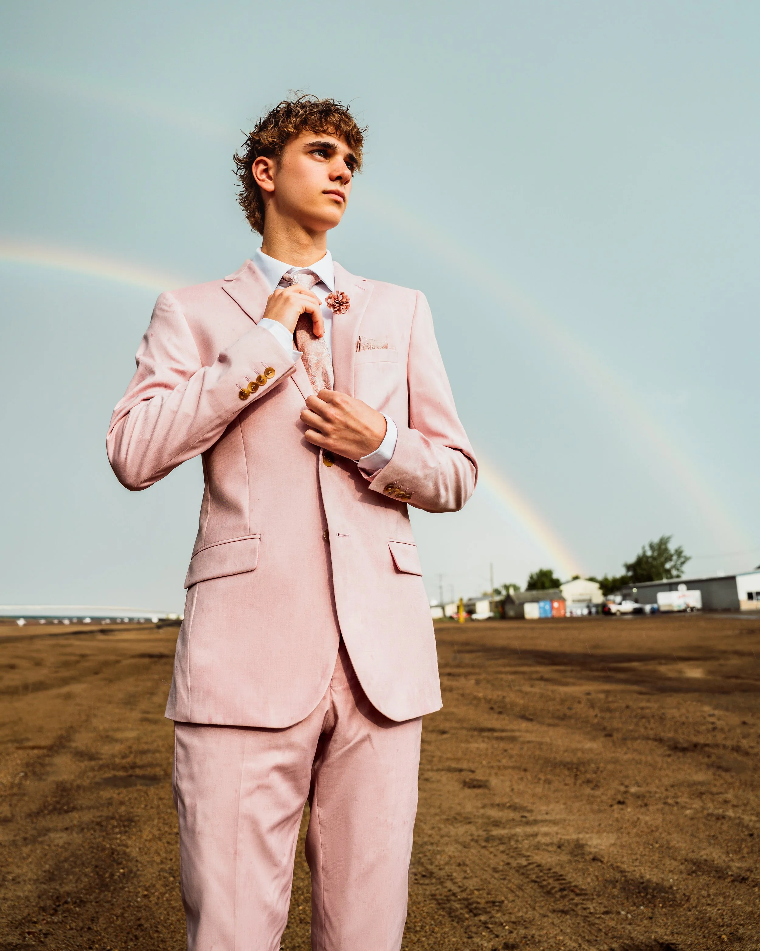 A young man wearing a pink suit adjusting his tie, standing outdoors with a rainbow in the cloudy sky behind him. By Canadian James Photography in Calgary, Alberta.
