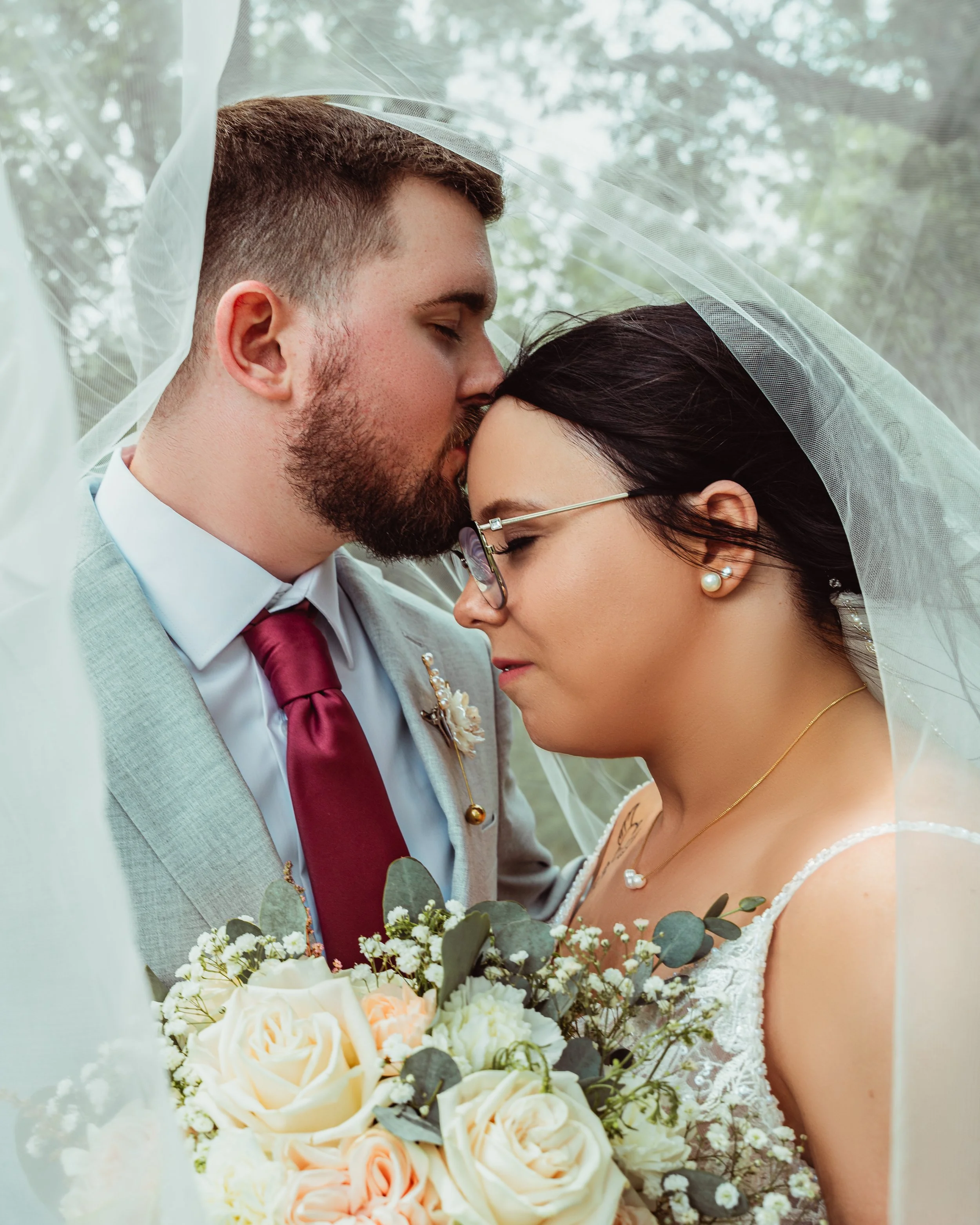A bride and groom, close-up, sharing a kiss under a veil during a wedding ceremony, with a bouquet of white and peach roses and greenery. Cochrane wedding photographer.