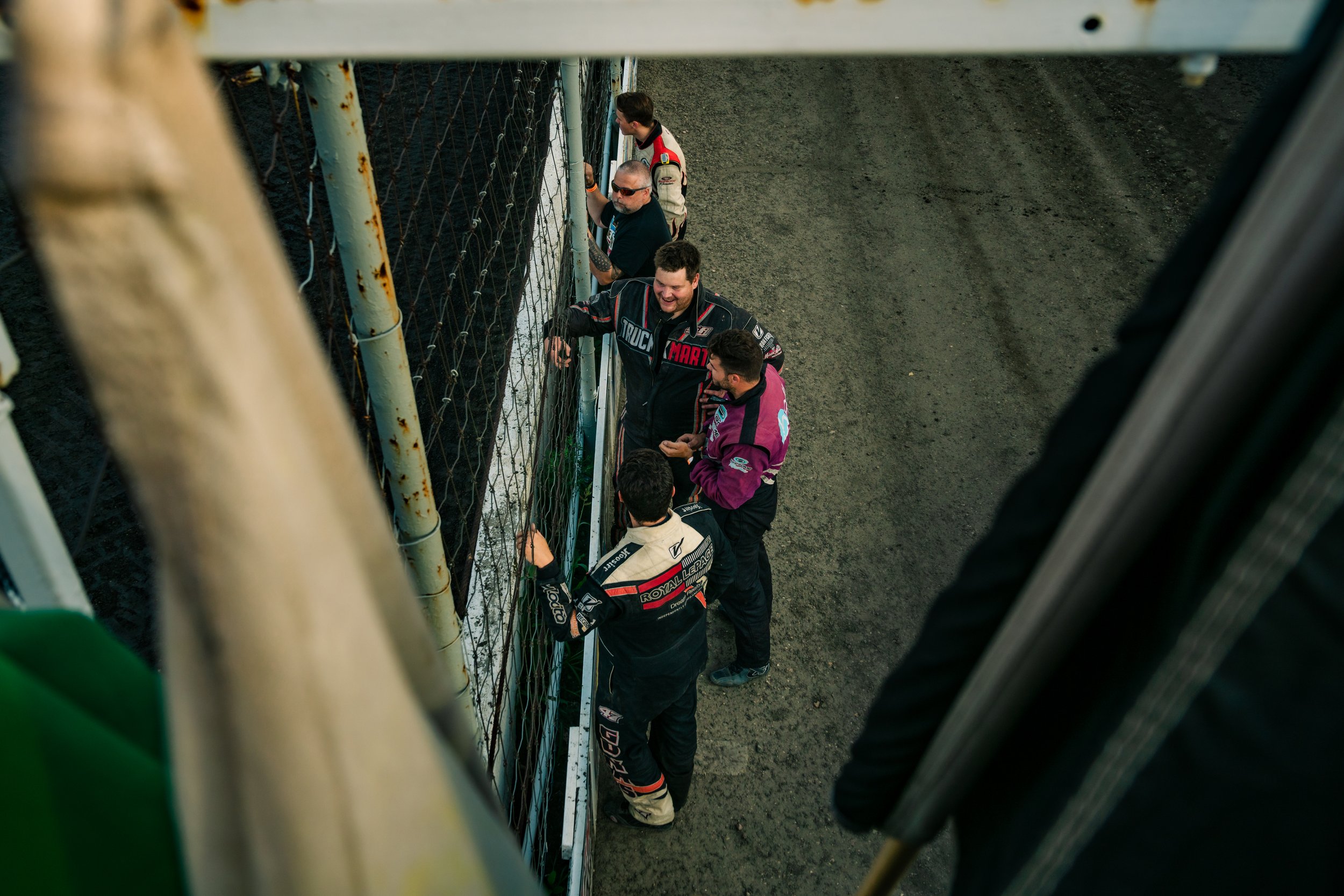 Group of race car drivers in suits standing by a fence, conversing at a racetrack. Canadian James Photography. Estevan, Saskatchewan.