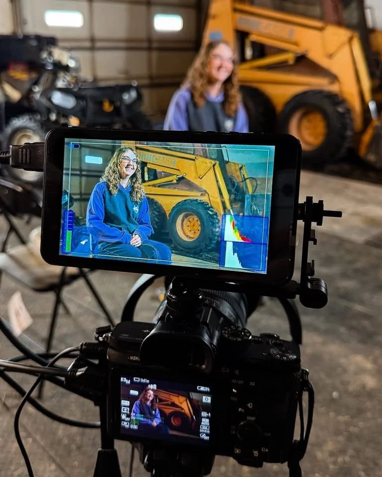 A woman with curly blonde hair and glasses sitting in front of construction equipment. Filming or photographing her with a camera setup inside a garage or warehouse.