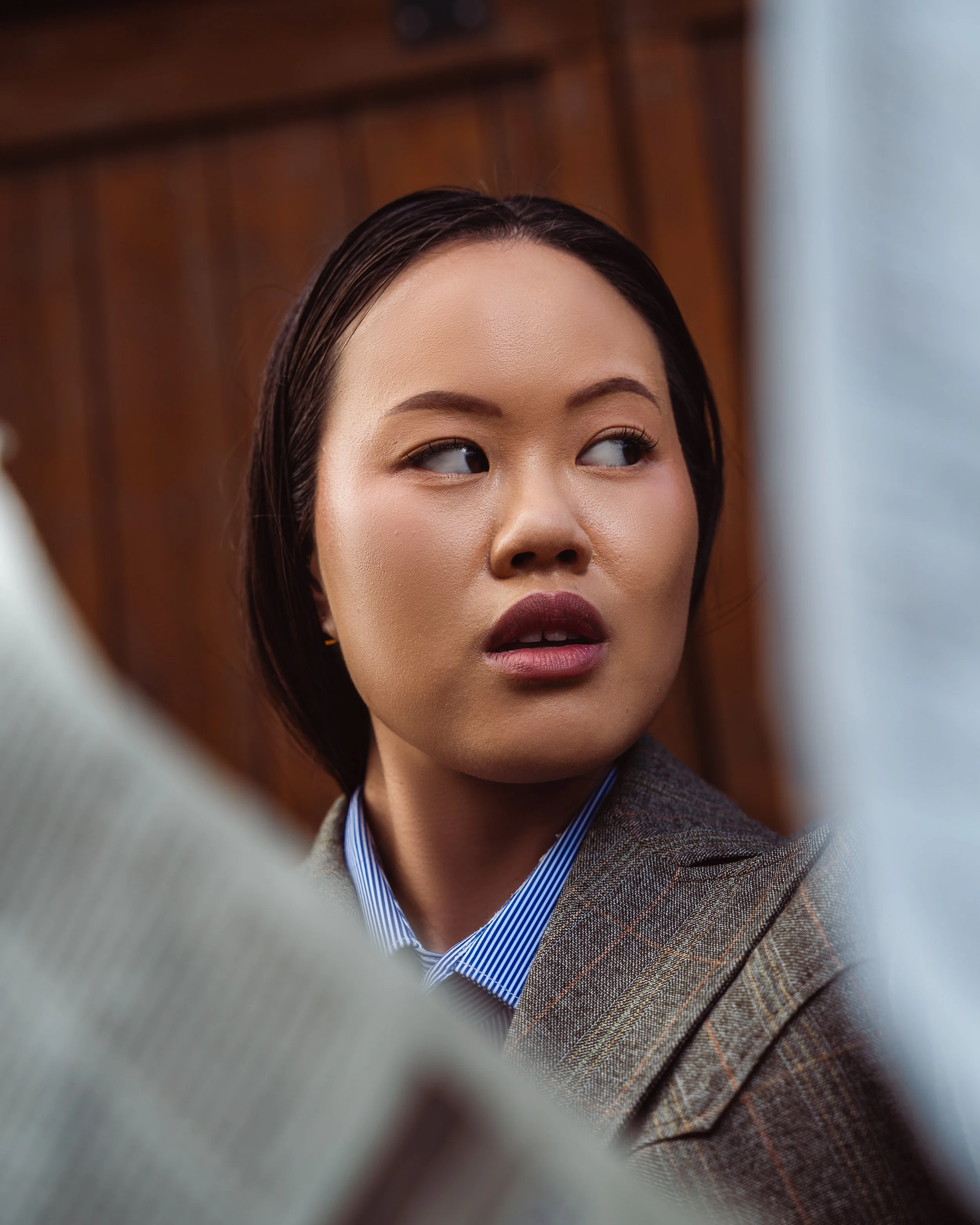 Close-up of a woman with dark hair, wearing a pinstripe suit and a blue-striped shirt, looking to her right, with a blurred wooden background. Fashion portraits in Cochrane, Alberta by Canadian James Photography.