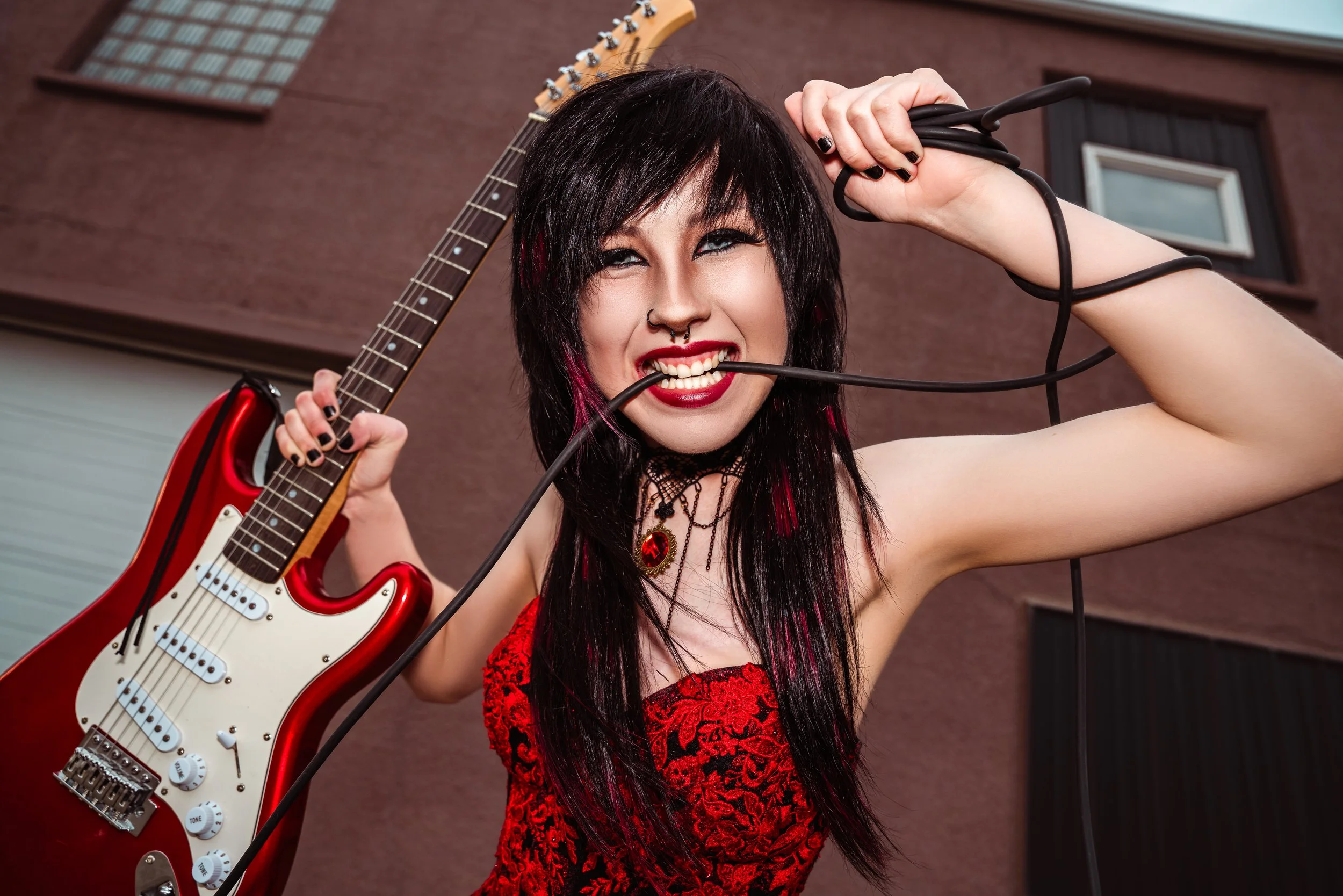 A woman with black hair and red lipstick holding a red and white electric guitar, smiling playfully with a cable in her mouth, standing outdoors in front of a brown building. By Canadian James Photography in Calgary, Alberta.