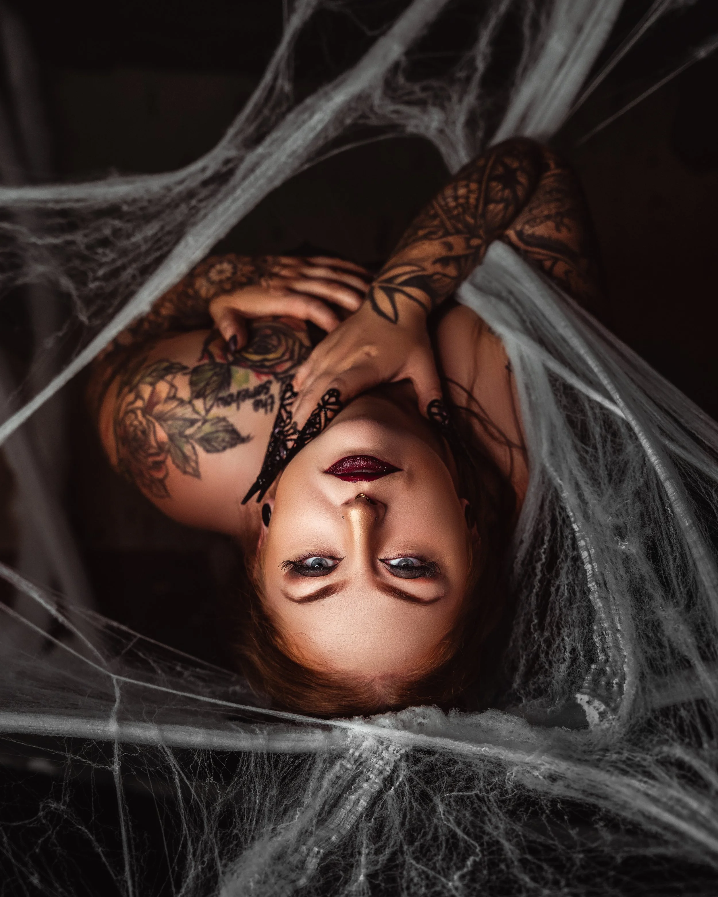 Woman with tattoos and dark makeup lying among cobwebs, looking up at camera