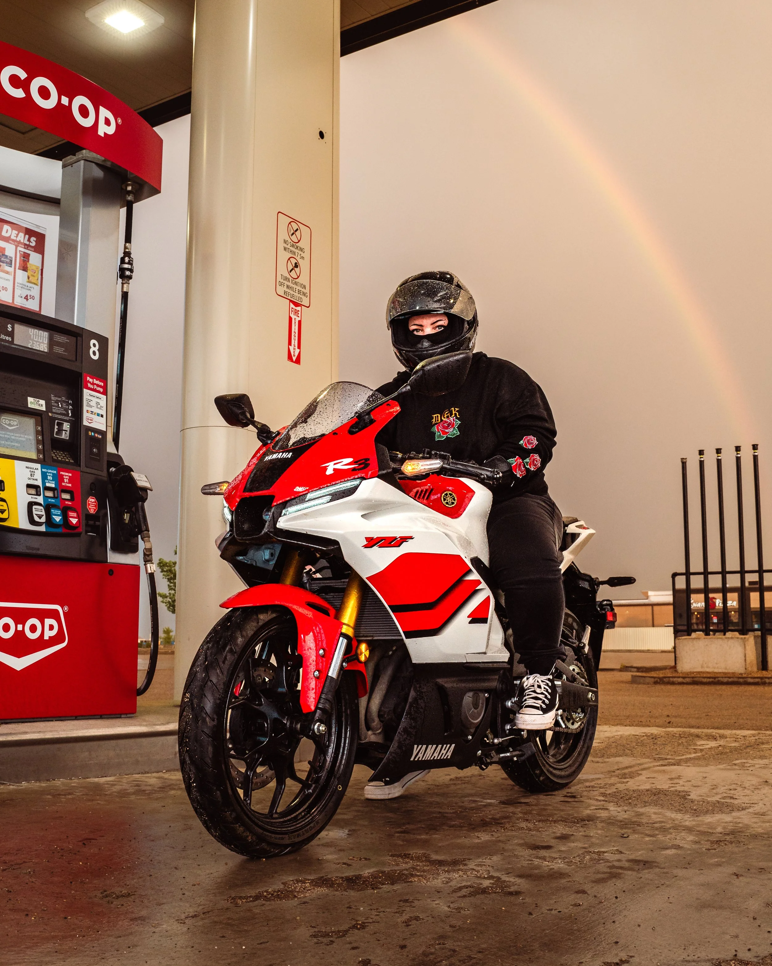 A person wearing a black helmet and black jacket sitting on a red and white Yamaha motorcycle at a gas station, with a rainbow visible in the sky in the background. By Canadian James Photography in Calgary, Alberta.