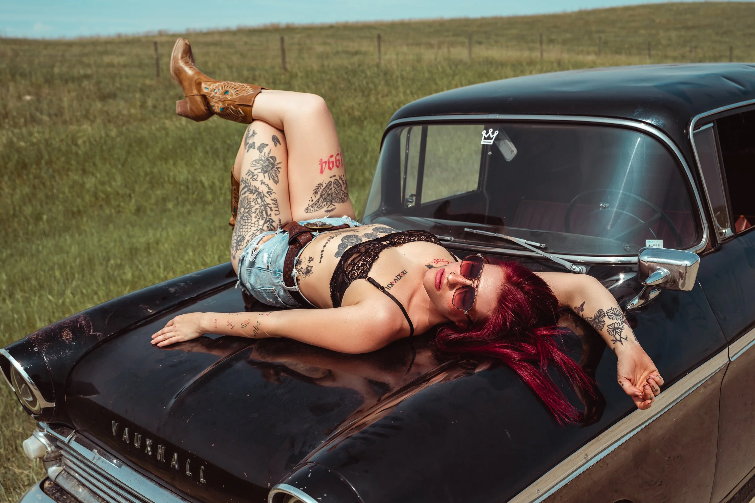 Woman with red hair and tattoos lying on the hood of a vintage black Vauxhall car in a grassy field, wearing sunglasses, a black lace top, and denim shorts.