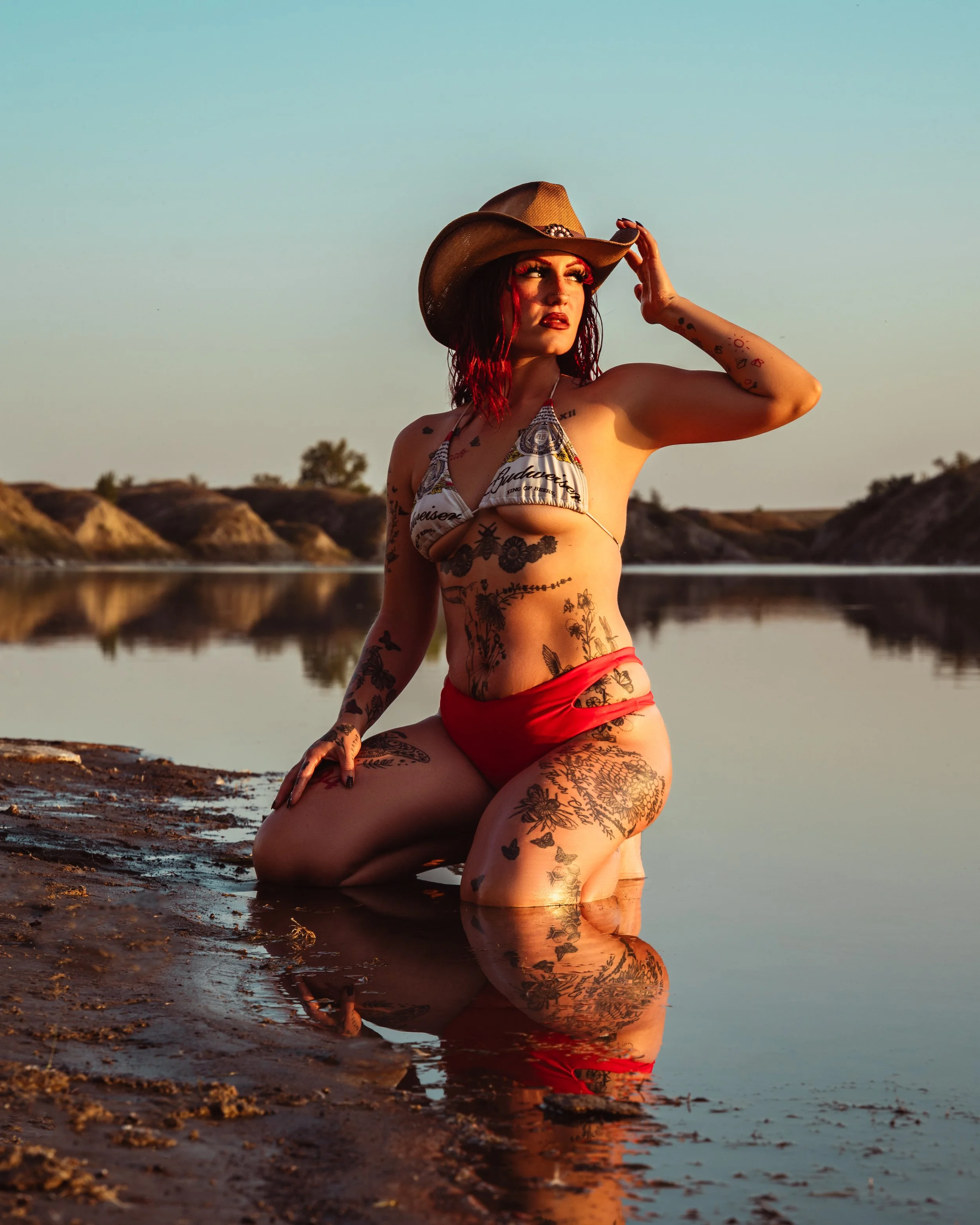 A tattooed woman with red hair wearing a bikini and cowboy hat kneeling by water at sunset, with reflections and distant hills in the background.