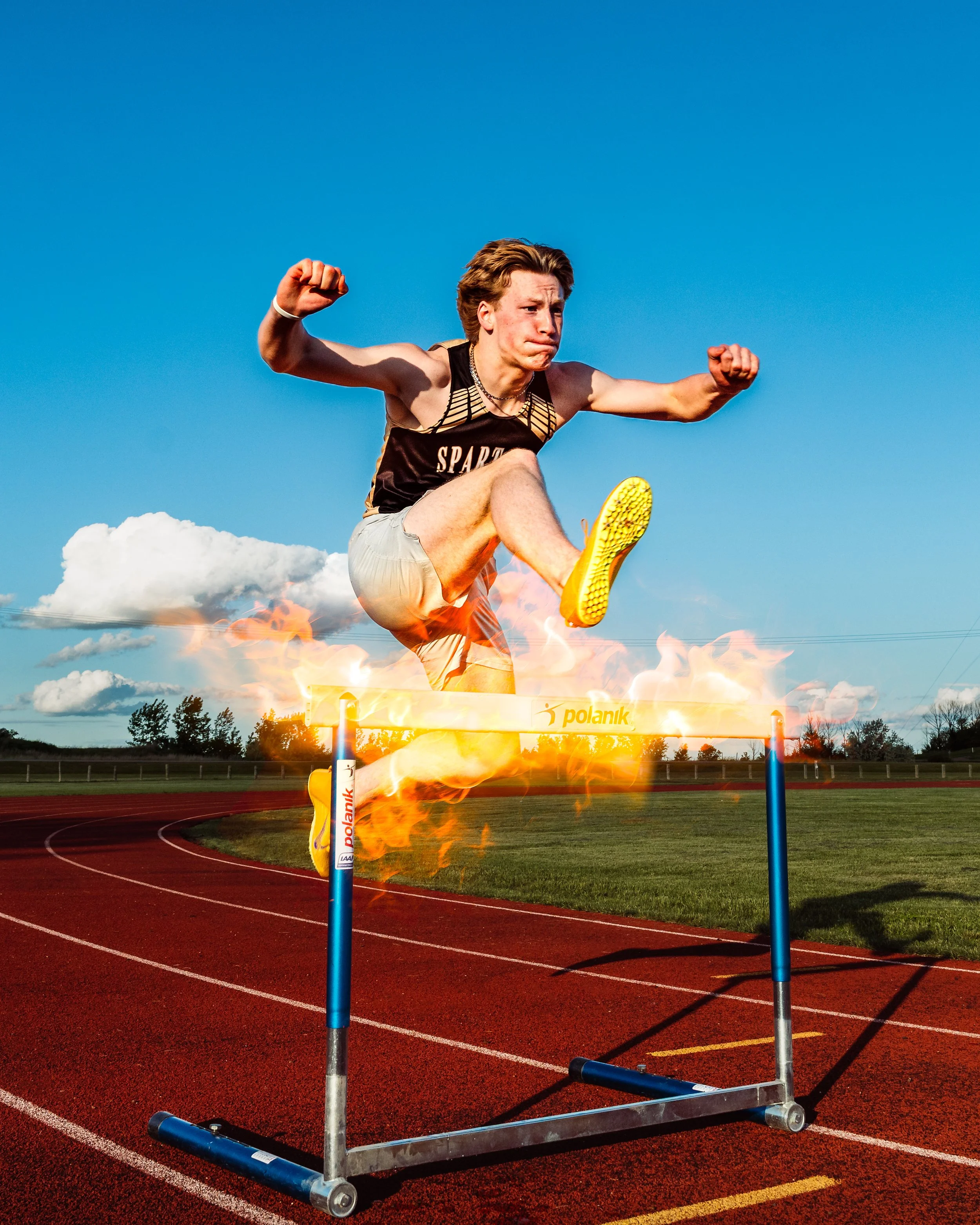 A young male athlete jumping over a hurdle on a running track during daytime, with a blue sky and clouds in the background. Cochrane Alberta photographer.