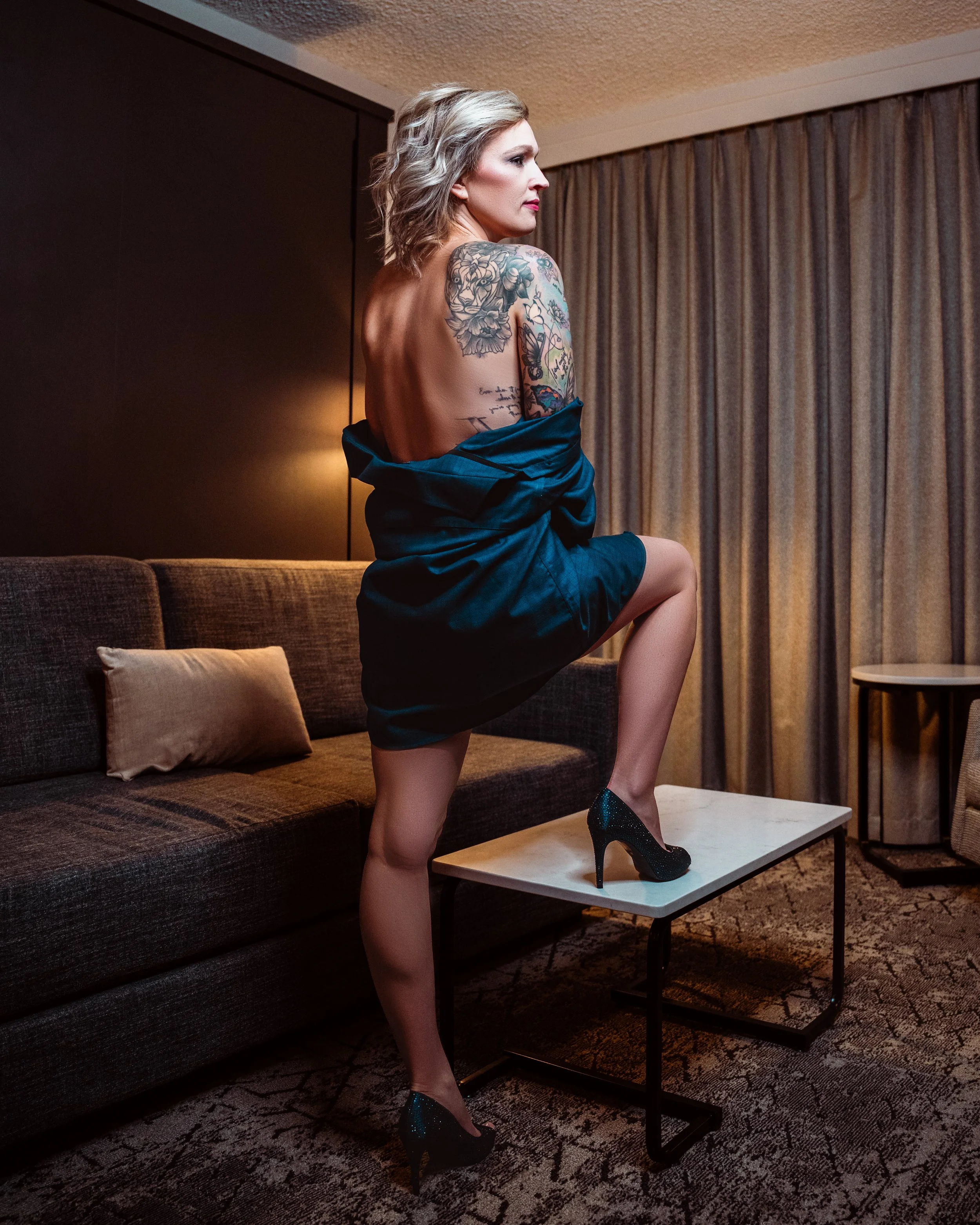 A woman with blonde hair and tattoos on her shoulder and arm, wearing high heels, sitting on a coffee table with one leg up, in a hotel room with a couch, pillow, and curtains.