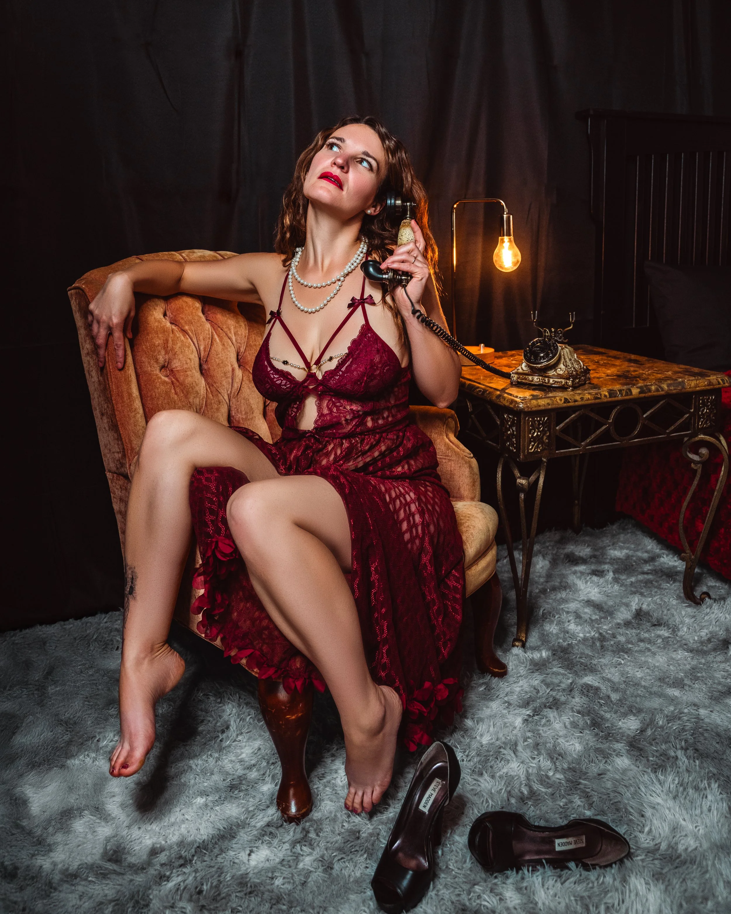 A woman in a red lingerie dress sitting on a vintage armchair, talking on an old-fashioned rotary phone, in a dimly lit room with a black backdrop, a side table with a lamp, and black high-heeled shoes on the floor.