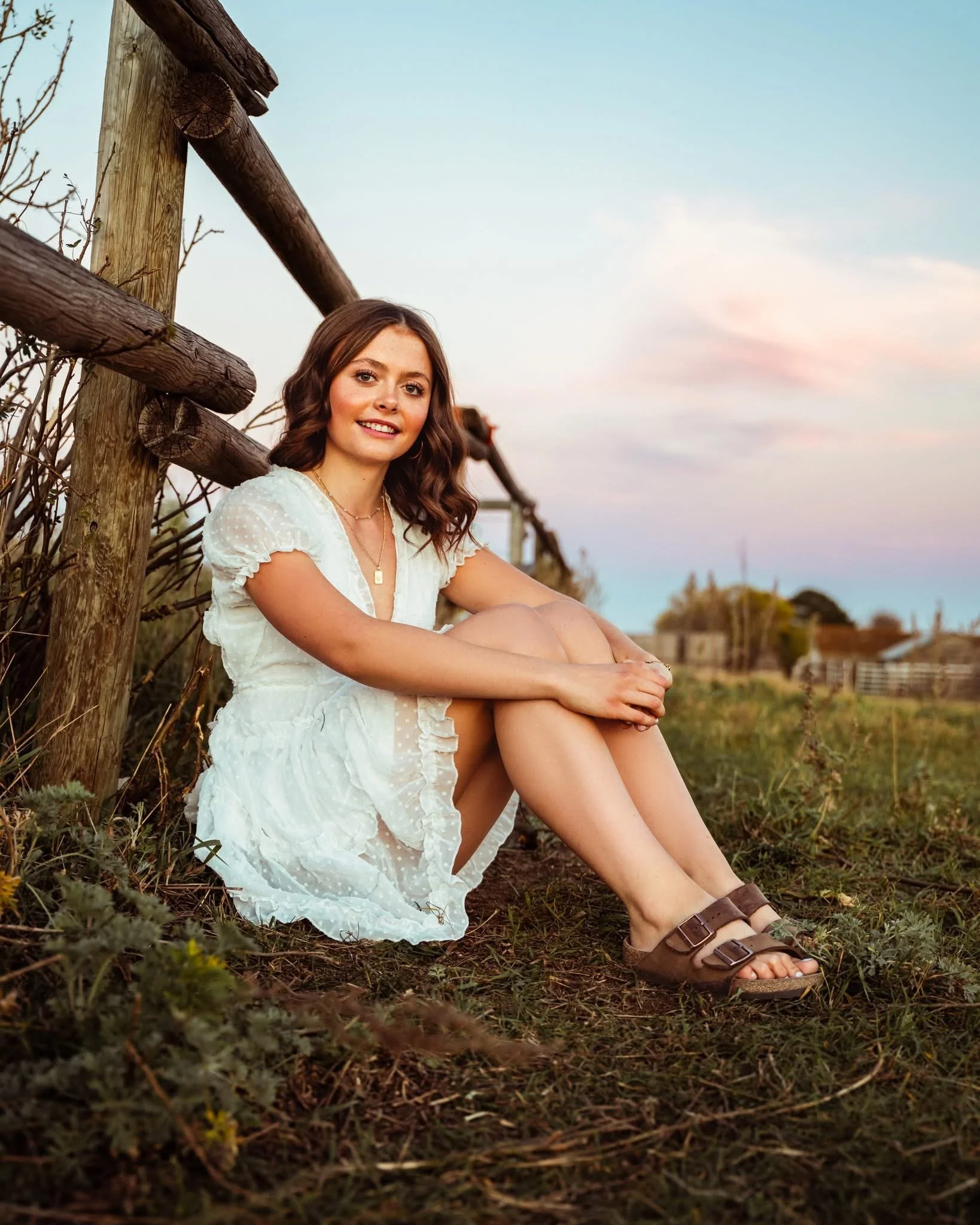 Young woman sitting outside near a rustic wooden fence during sunset, wearing a white dress and sandals, with a smile and relaxed pose. Cochrane, Alberta grad photographer.
