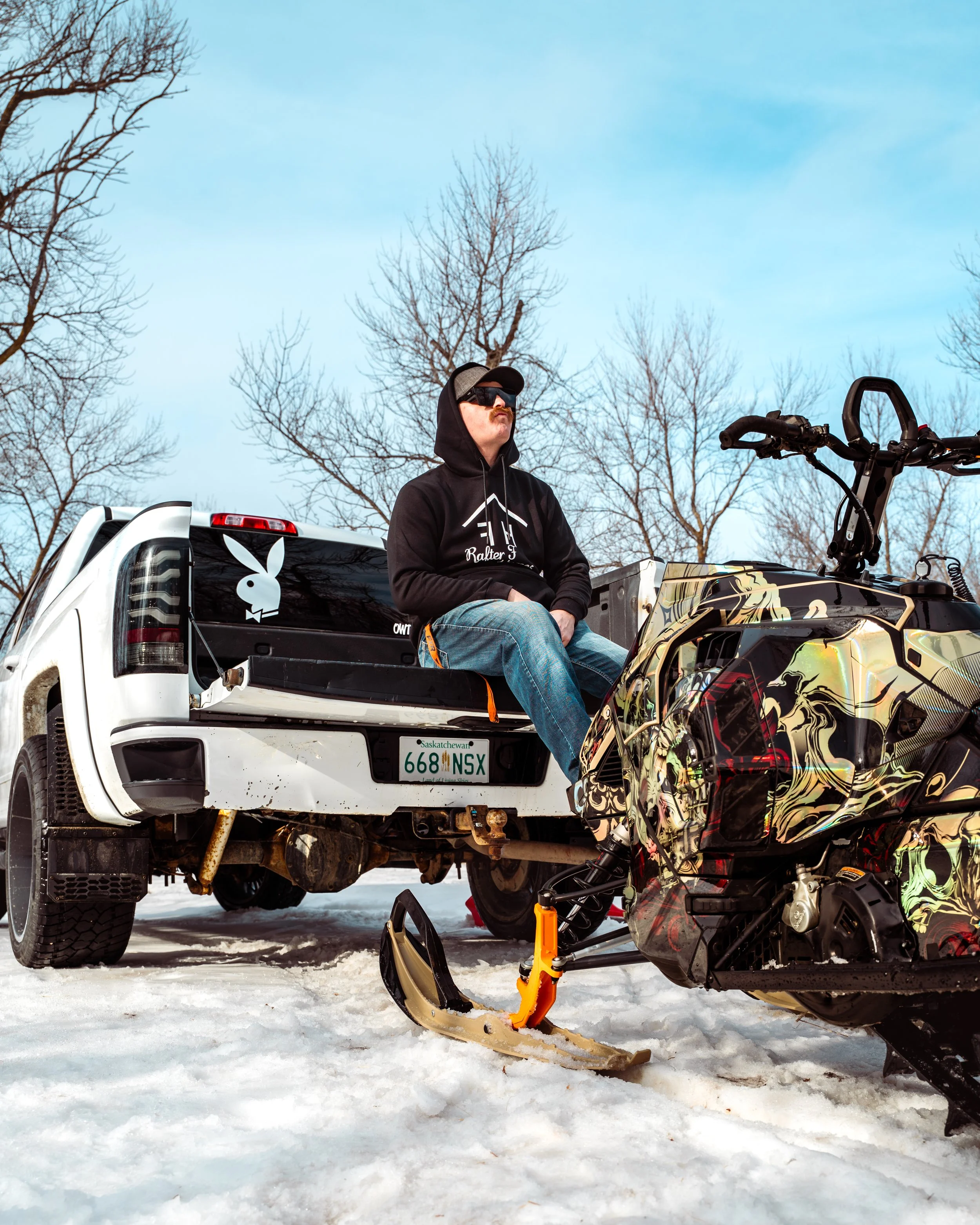 A man sitting on the tailgate of a white pickup truck, wearing a black hoodie, jeans, sunglasses, and a cap, outdoors in a snowy area with bare trees. A snowmobile with a colorful design is in the foreground.