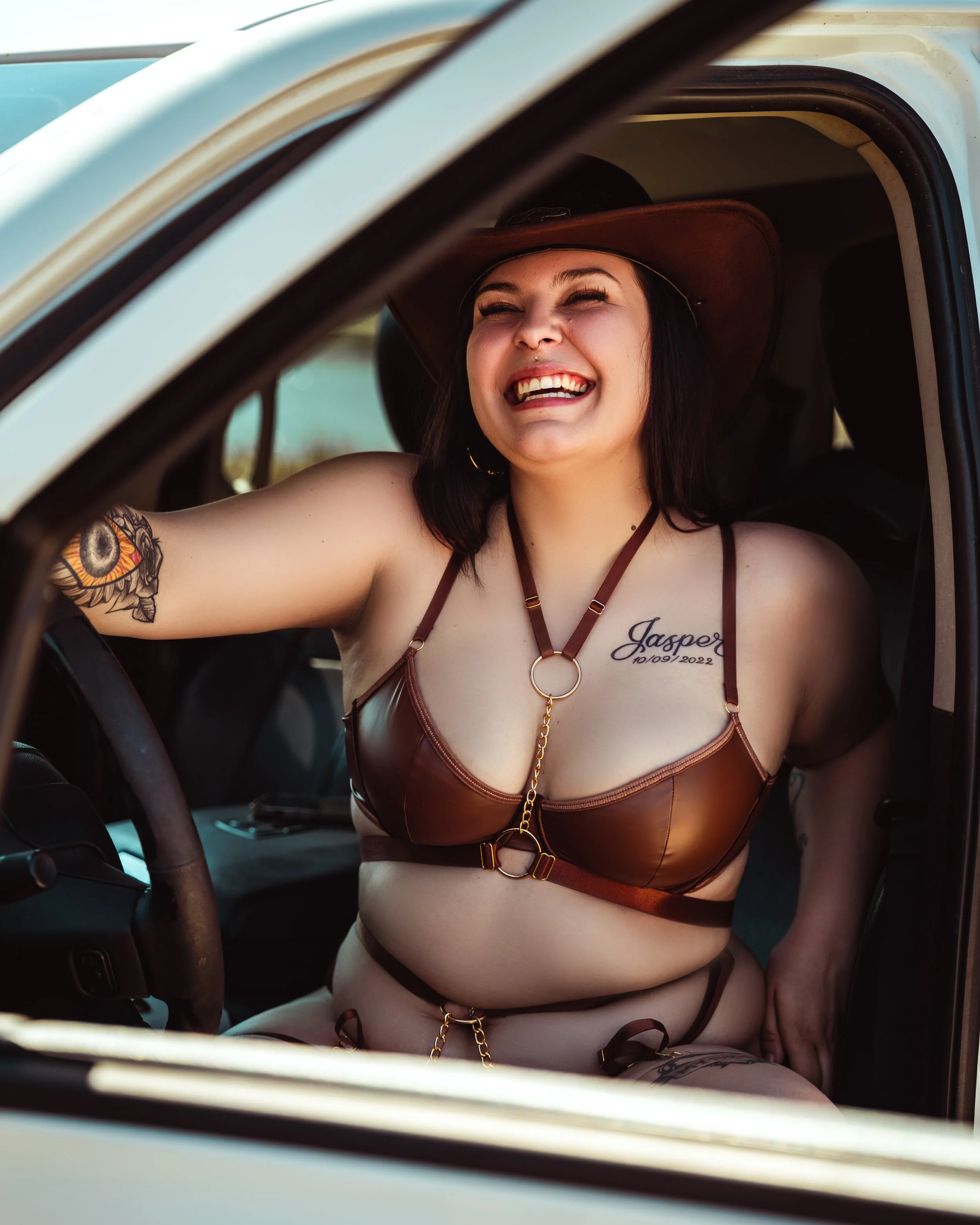 A woman named Jasper sitting in a vehicle, wearing a leather harness and a wide-brimmed hat, smiling happily while looking outside.