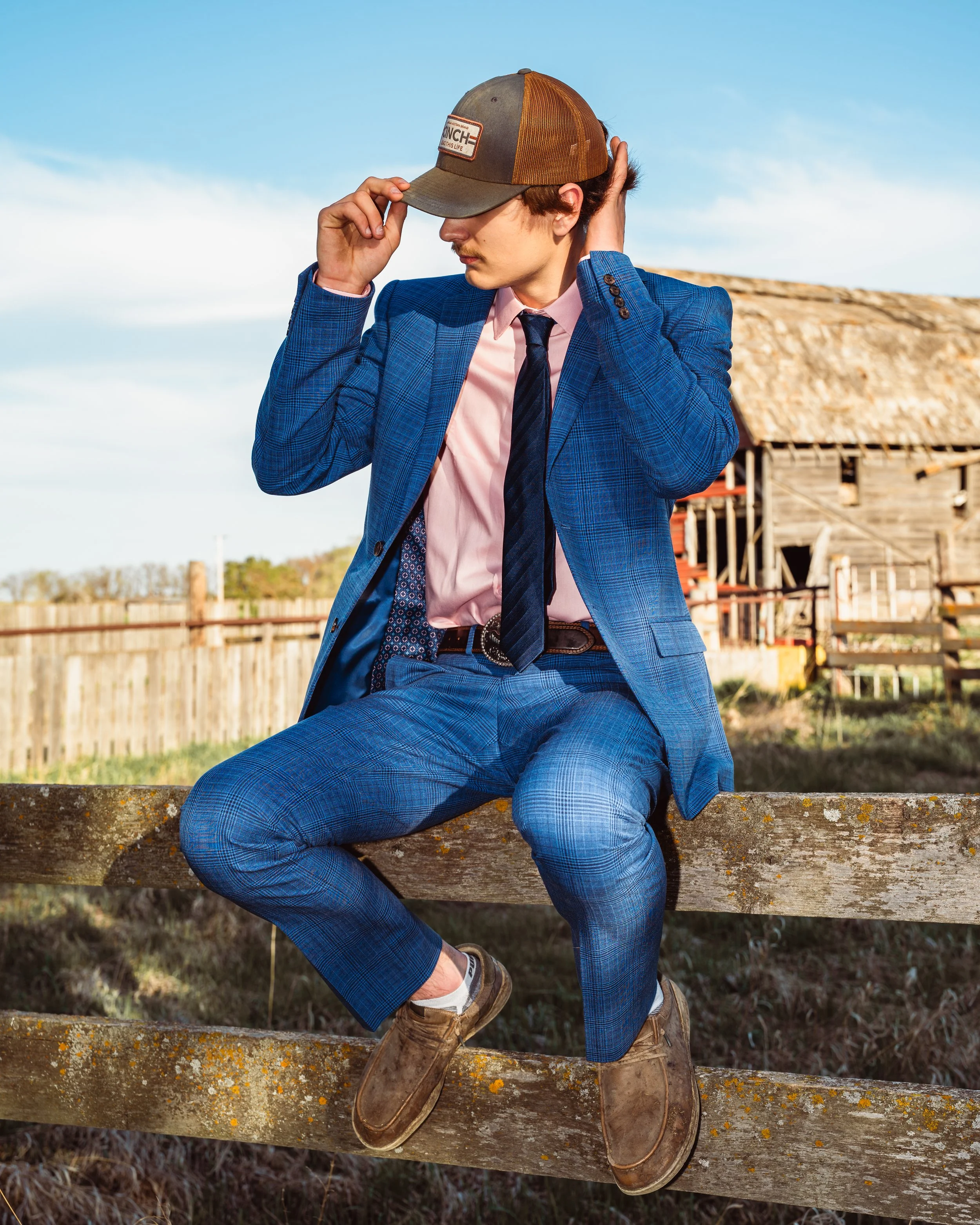 A young man dressed in a blue checkered suit, pink dress shirt, and dark tie, sitting on a wooden fence with his right knee on the fence and his left foot on the ground. He is wearing brown shoes and a gray cap, adjusting his cap with his right hand.