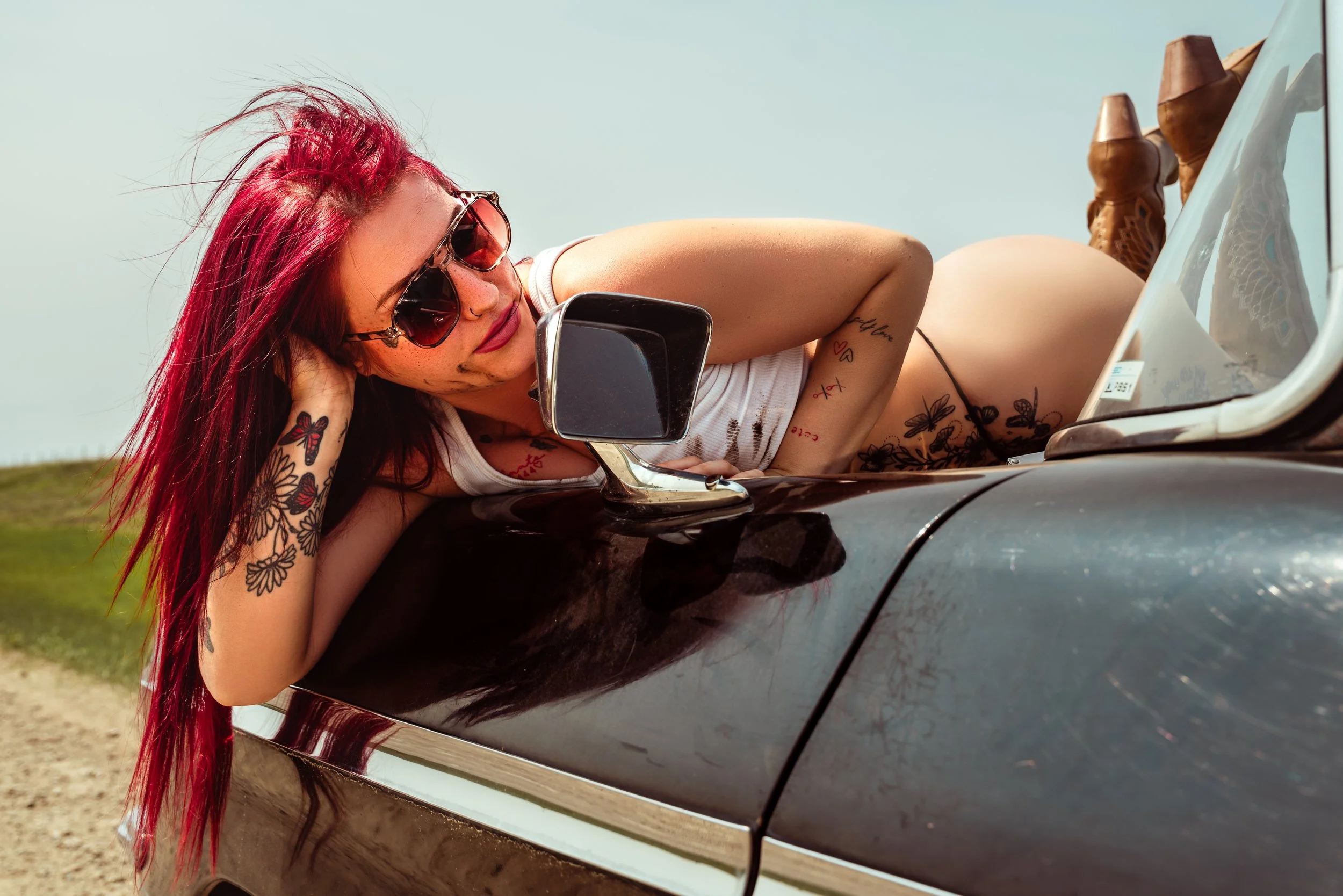 A woman with red hair and tattoos lying on the side of a vintage car with sunglasses, wearing a white tank top and black underwear, outdoors on a sunny day.