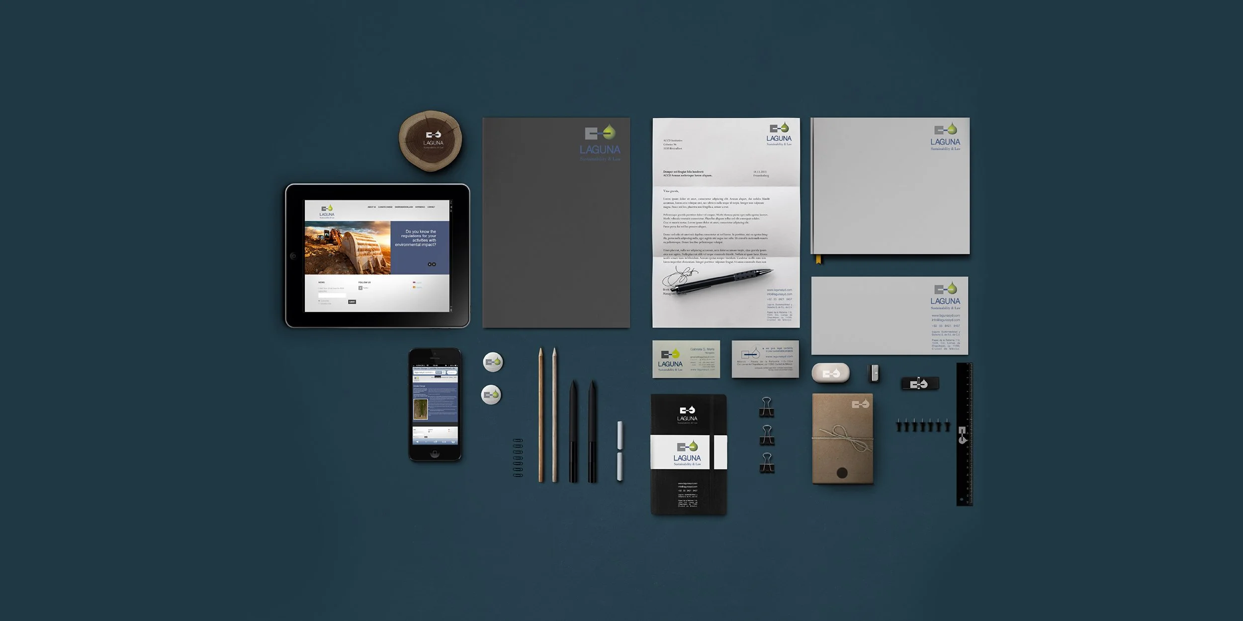 A flat lay of corporate branding materials for Laguna, including a tablet, smartphone, business cards, letterhead, notebooks, pens, erasers, push pins, and other office supplies, all arranged on a dark teal surface.