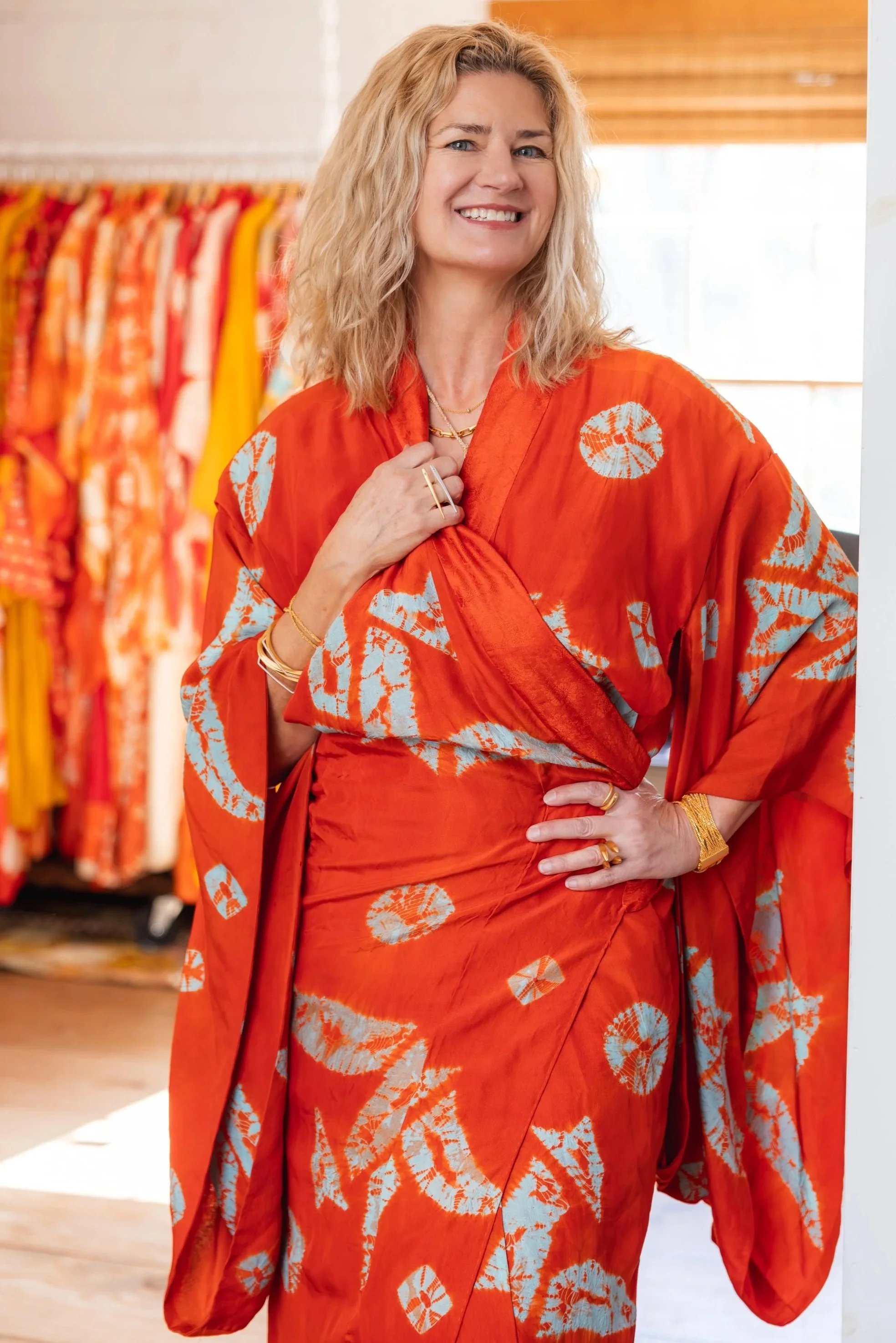 Vandi Hodges smiling, wearing a bright orange dress and kimono with white and blue circular patterns, standing in a clothing store with colorful garments hanging in the background.