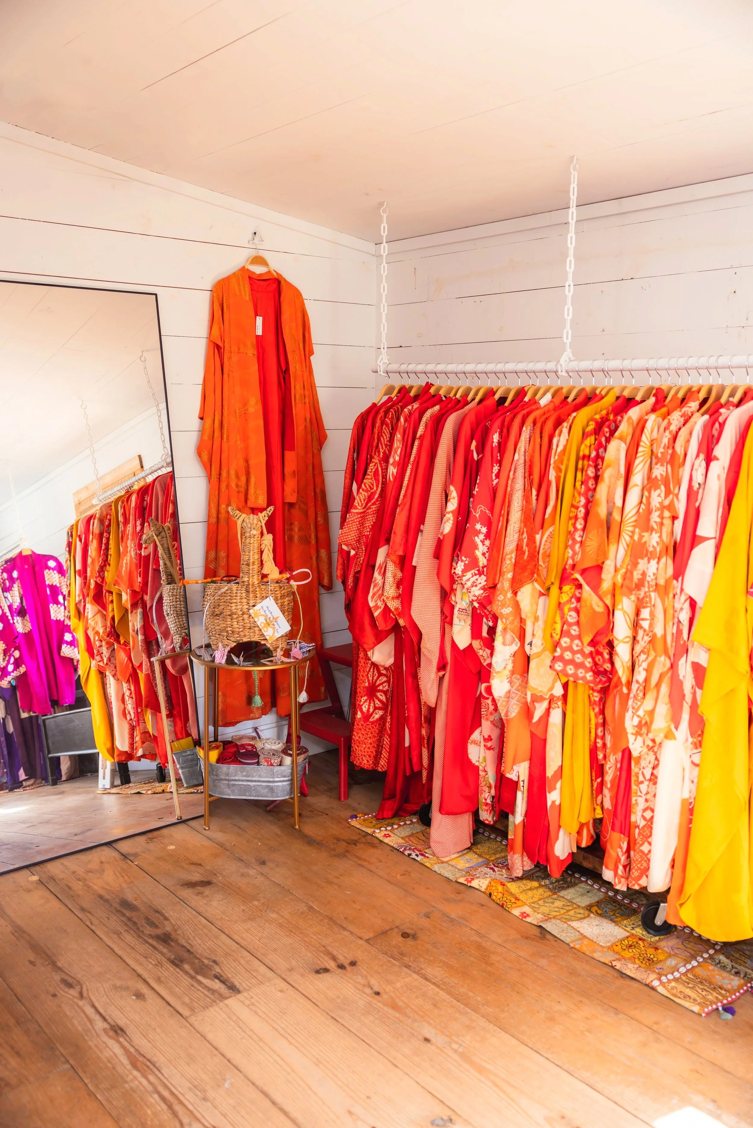 A clothing store display with red, orange, yellow, and pink kimonos on a rack, a tall mirror, and a small table with woven baskets and accessories on a wooden floor.