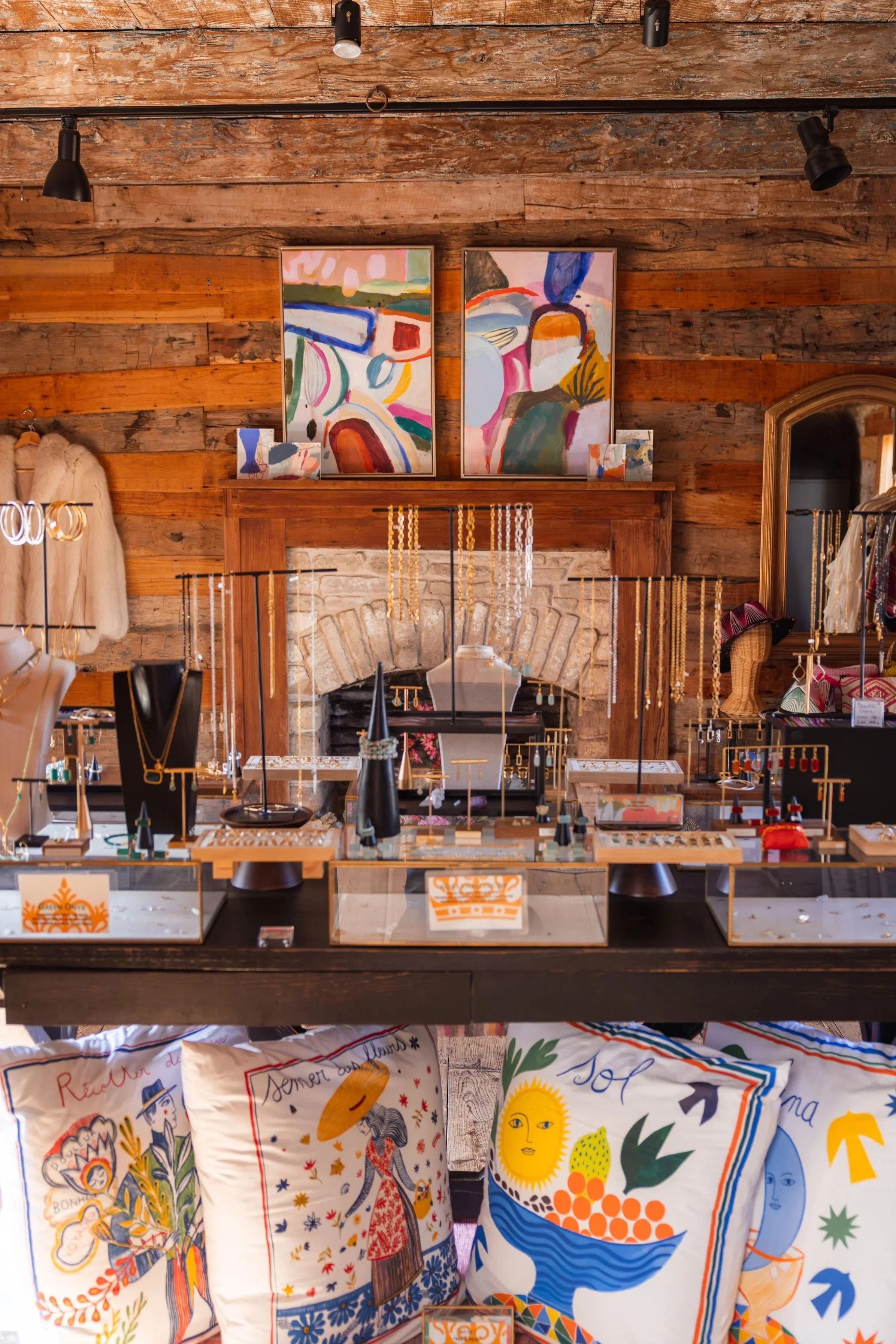 A retail store display with colorful merchandise, abstract paintings on a wooden wall, clothing items, jewelry, and decorative pillows with vibrant designs. Via Vandi in Round Top, Texas at Henkel Square.