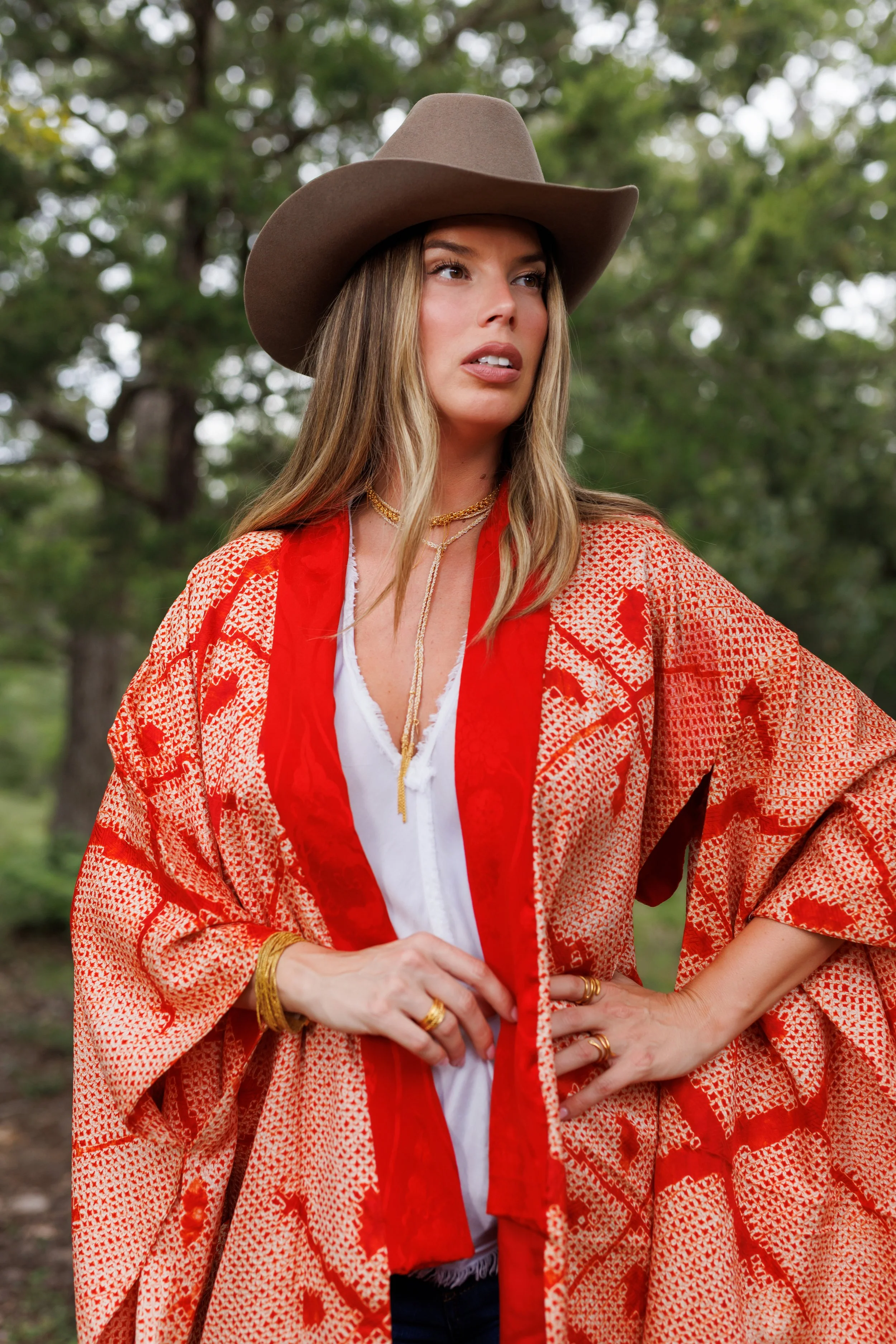 A woman wearing a large brown cowboy hat, a red patterned kimono, a white top, and gold jewelry by Via Vandi, standing outdoors with green trees in the background.