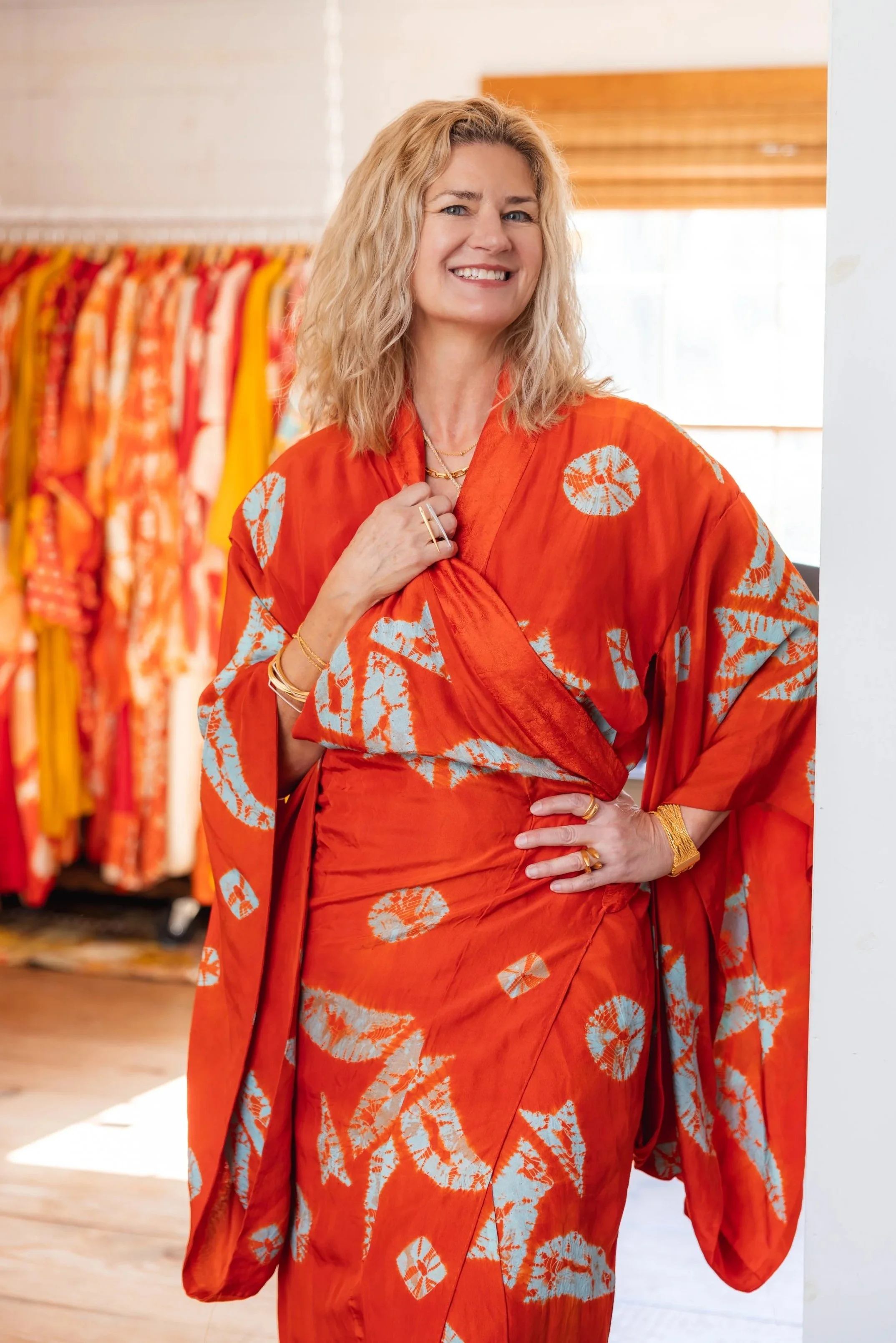 Vandi Hodges wearing a vibrant orange patterned kimono and matching dress, smiling, standing in front of a clothing rack filled with similar colorful garments in a store or boutique.