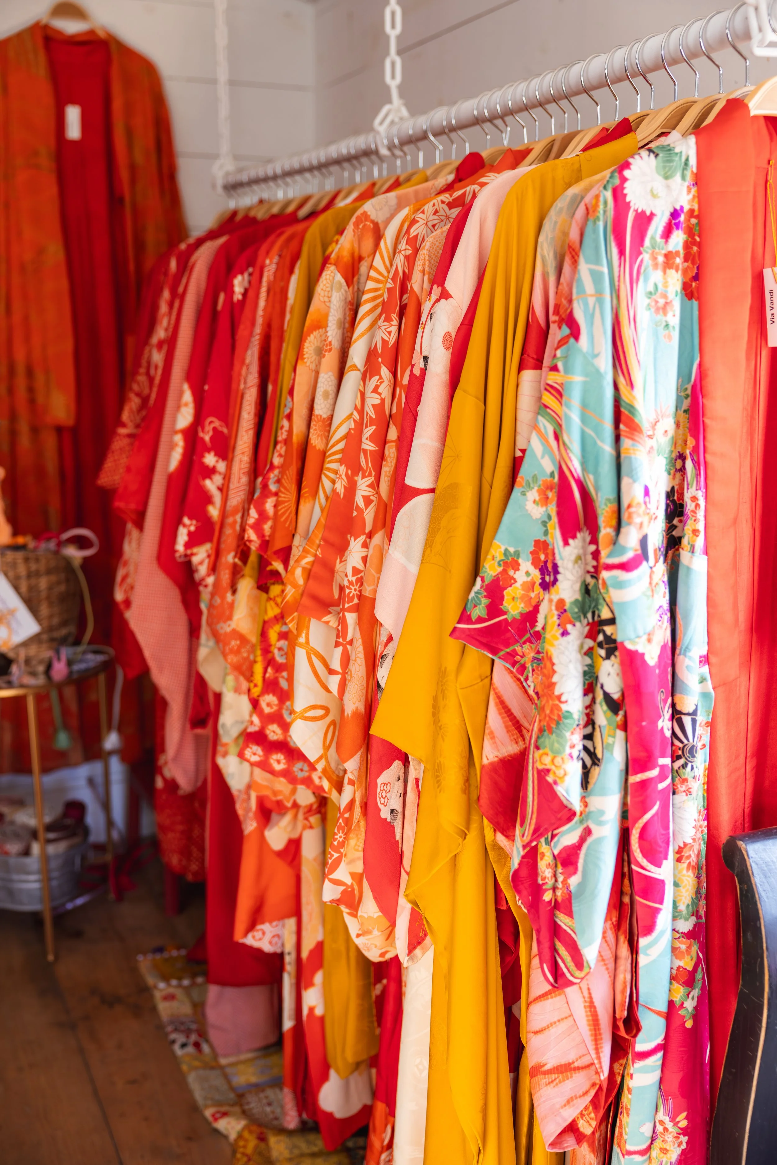 A clothing rack displays colorful floral and patterned kimonos in shades of red, orange, yellow, pink, blue, and green. The kimonos are hanging on wooden and metal hangers inside a boutique or clothing store.