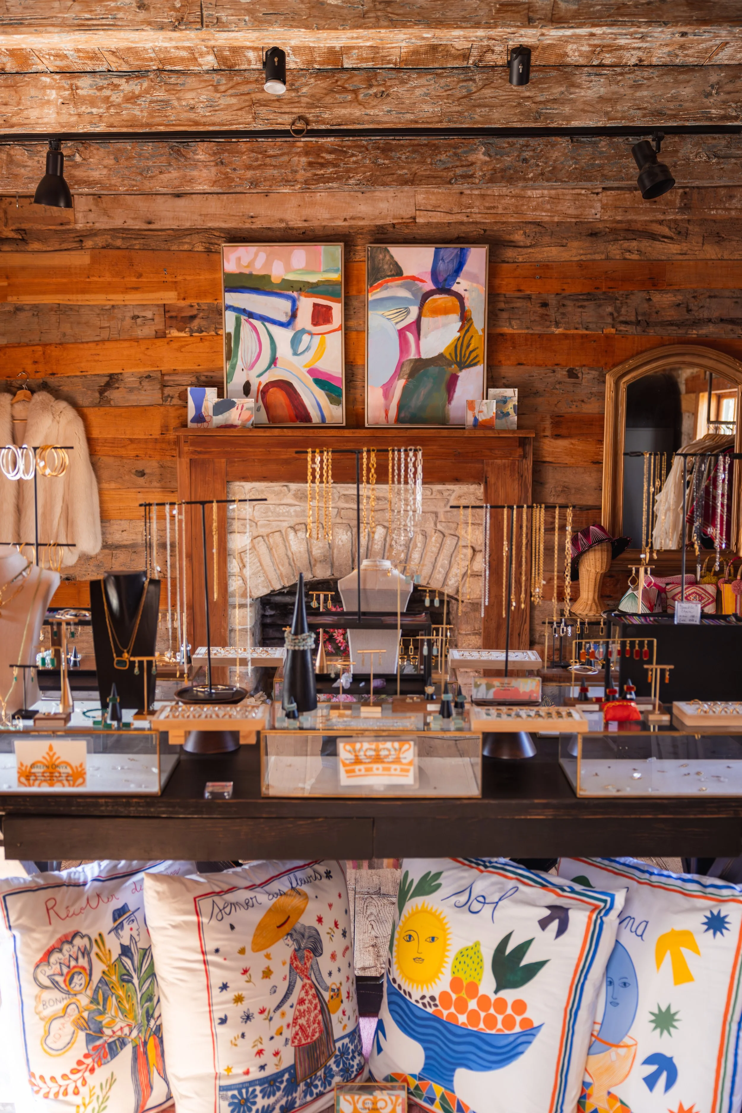 An interior of Via Vandi in Round Top, Texas at historic Henkel Square featuring a brick fireplace, colorful abstract paintings, jewelry displayed on stands, and printed tote bags with vibrant, whimsical designs.