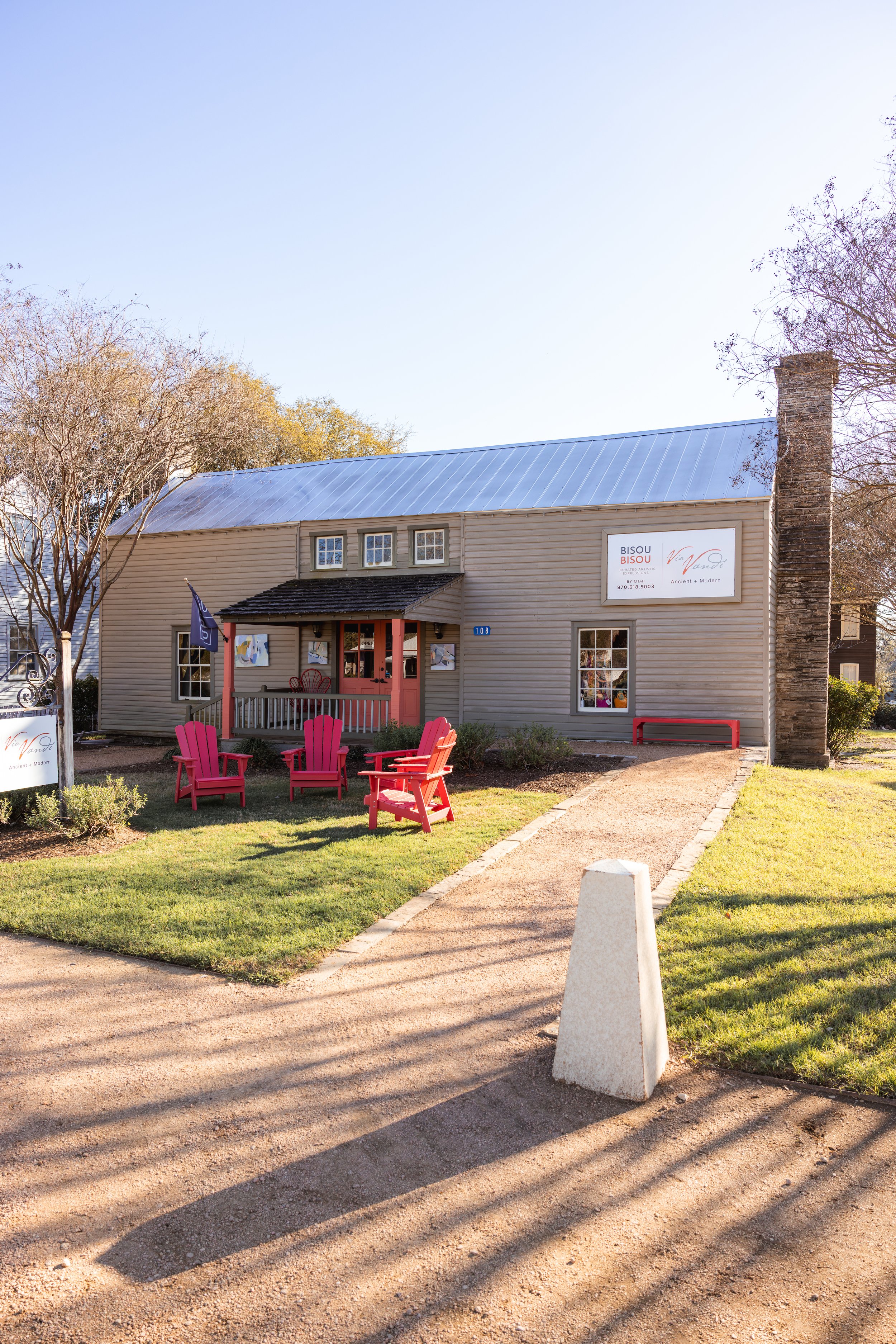 Front view of a gray wooden building with a metal roof, pink door, and outdoor seating, located on a grassy area with a dirt pathway and trees, sunlight casting shadows. Via Vandi in Round Top, Texas at Henkel Square.