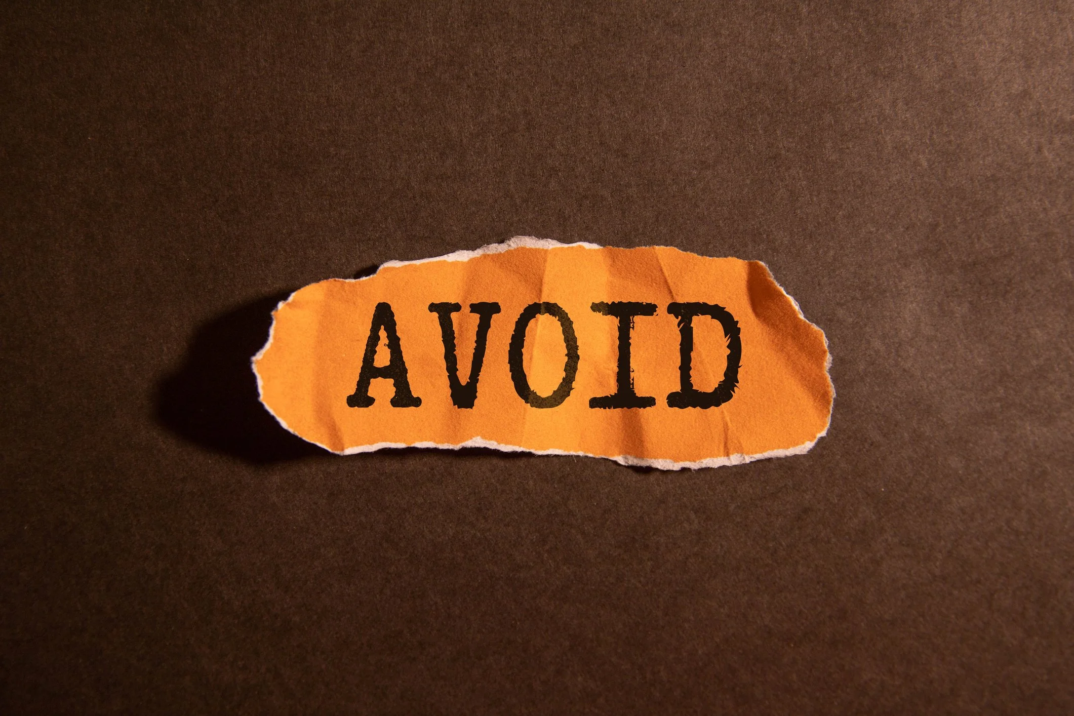 A torn piece of orange paper with the word 'AVOID' printed in black, placed on a dark brown surface with a shadow cast on one side.