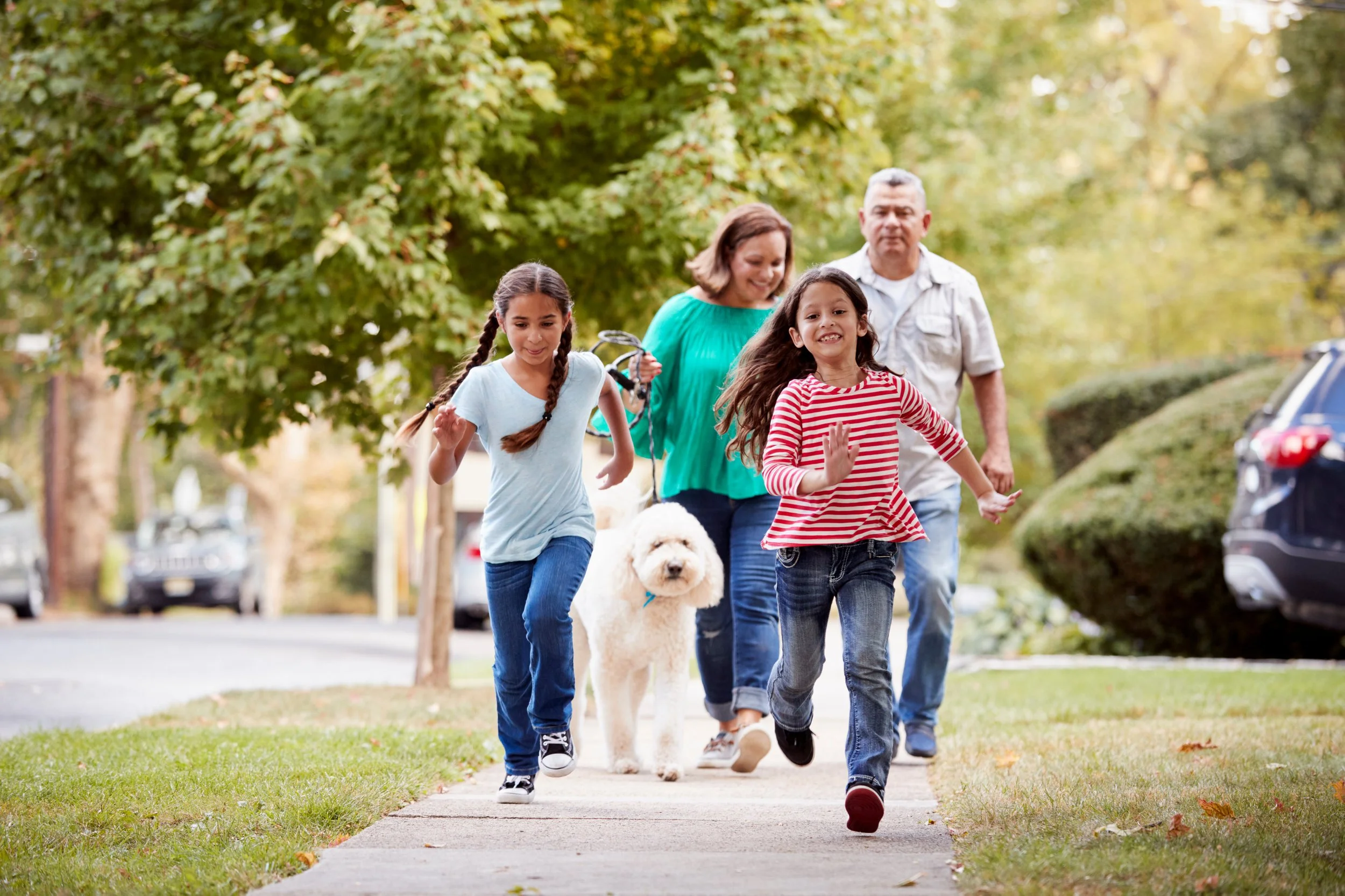 Family of four walking on a sidewalk, with two young girls in the front and their parents behind, accompanied by a white fluffy dog during daytime in a green, leafy neighborhood.