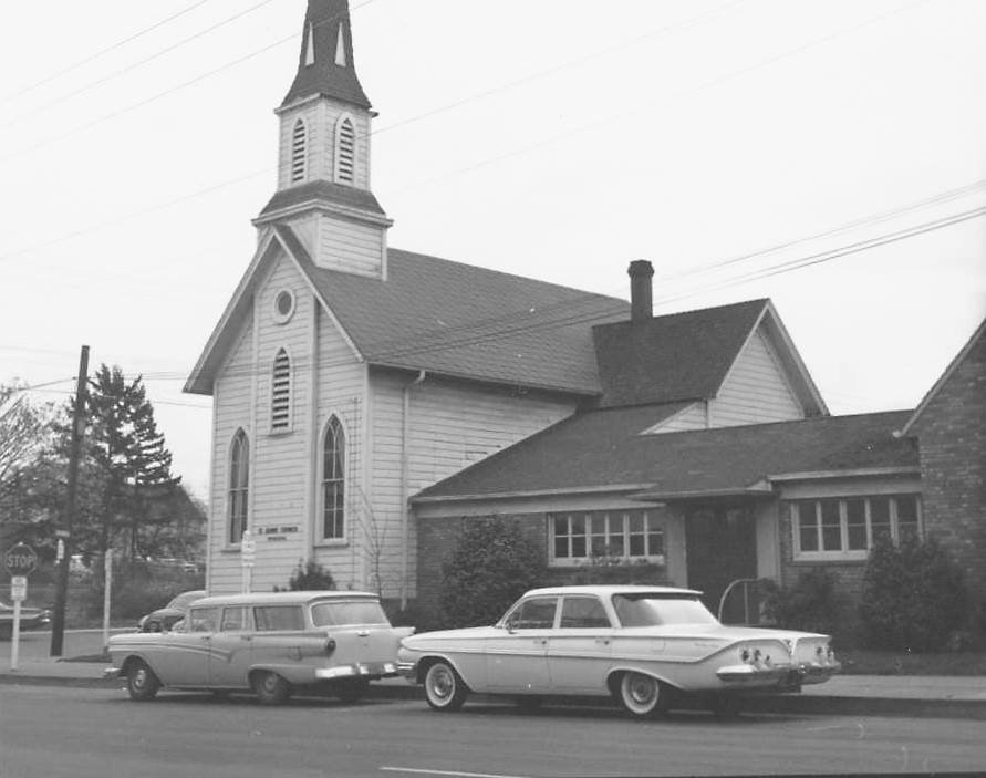 Little white church with a steeple with two mid-20th century cars parked in front